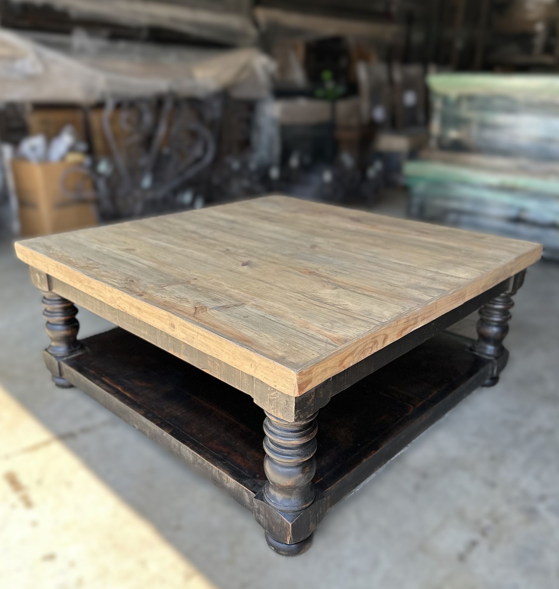 Square wooden coffee table with a light-colored top and dark, carved base and shelf. Background is a workshop or storage space.