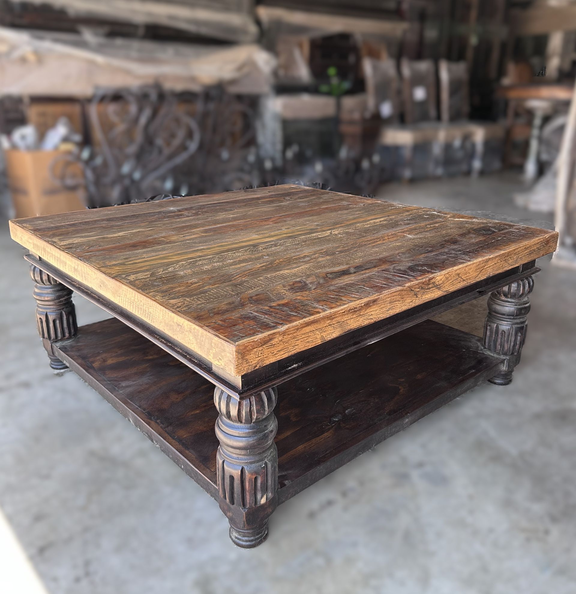 Square wooden coffee table with a shelf, in a warehouse setting. The table has ornate legs and a distressed wood finish.