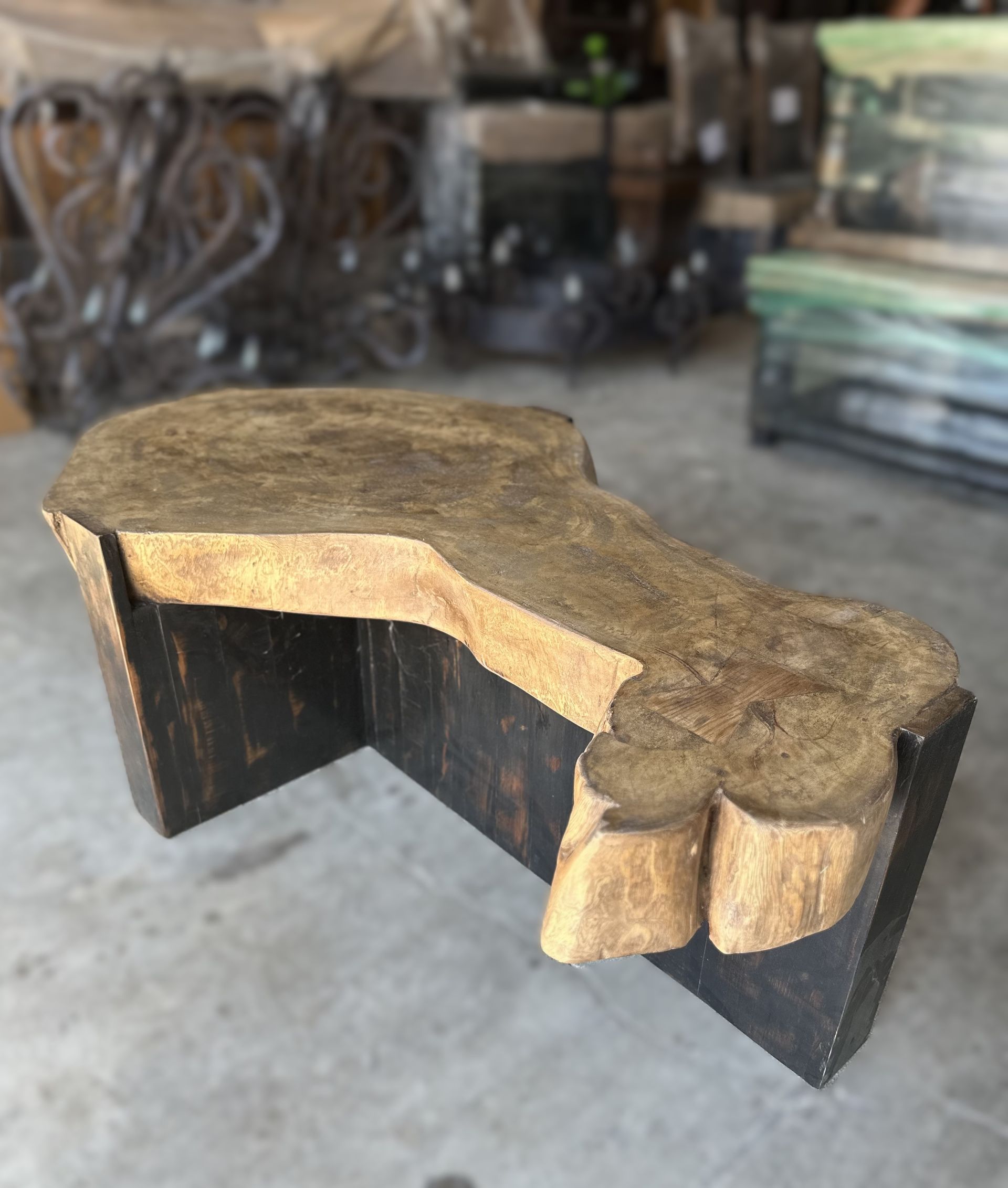 A rustic wooden bench with a natural edge on the top, featuring dark stained legs. Indoors on a concrete floor.