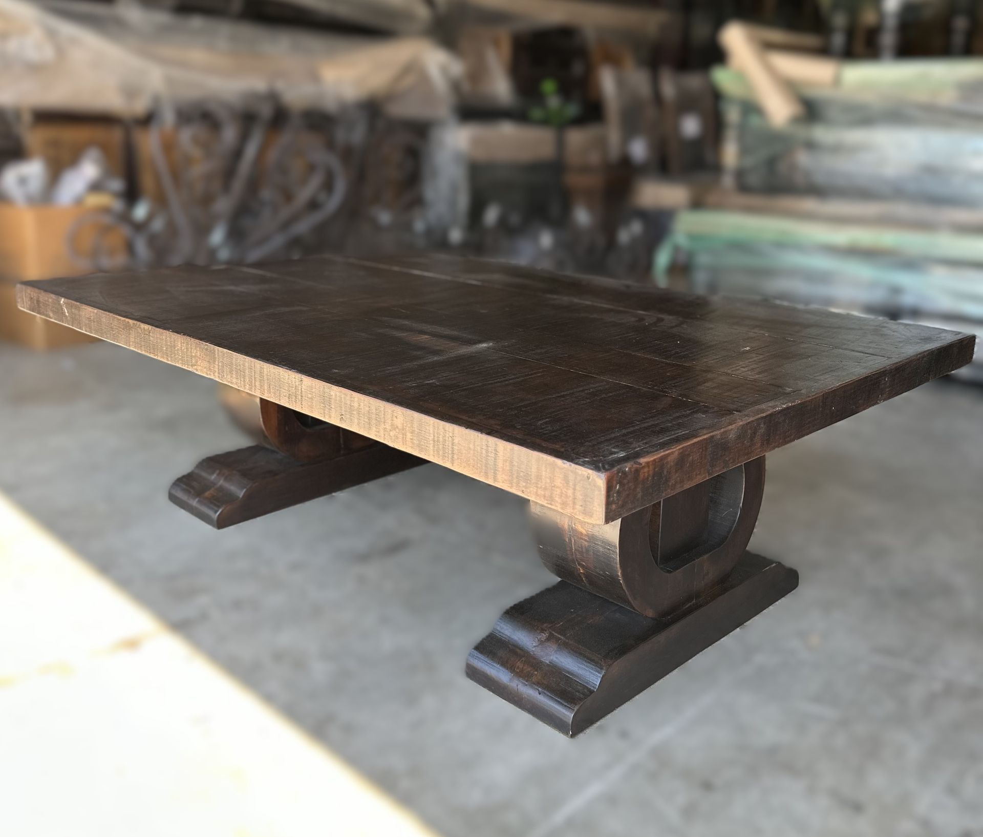 Dark brown wooden dining table with a thick rectangular top and two pedestal-style bases, set in a warehouse or workshop.
