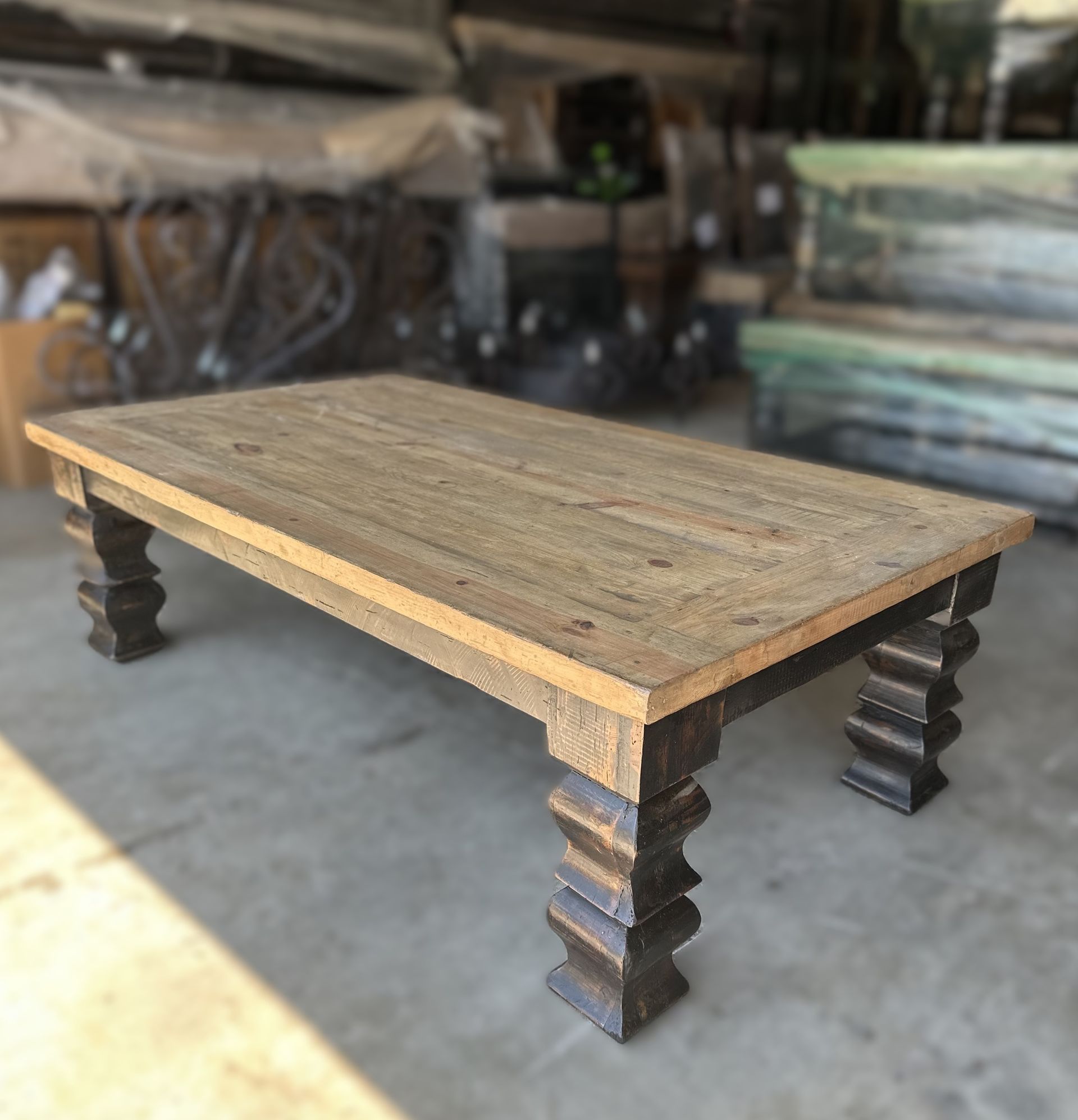 A rustic wooden coffee table with dark, carved legs stands in a warehouse. The table has a light brown top.