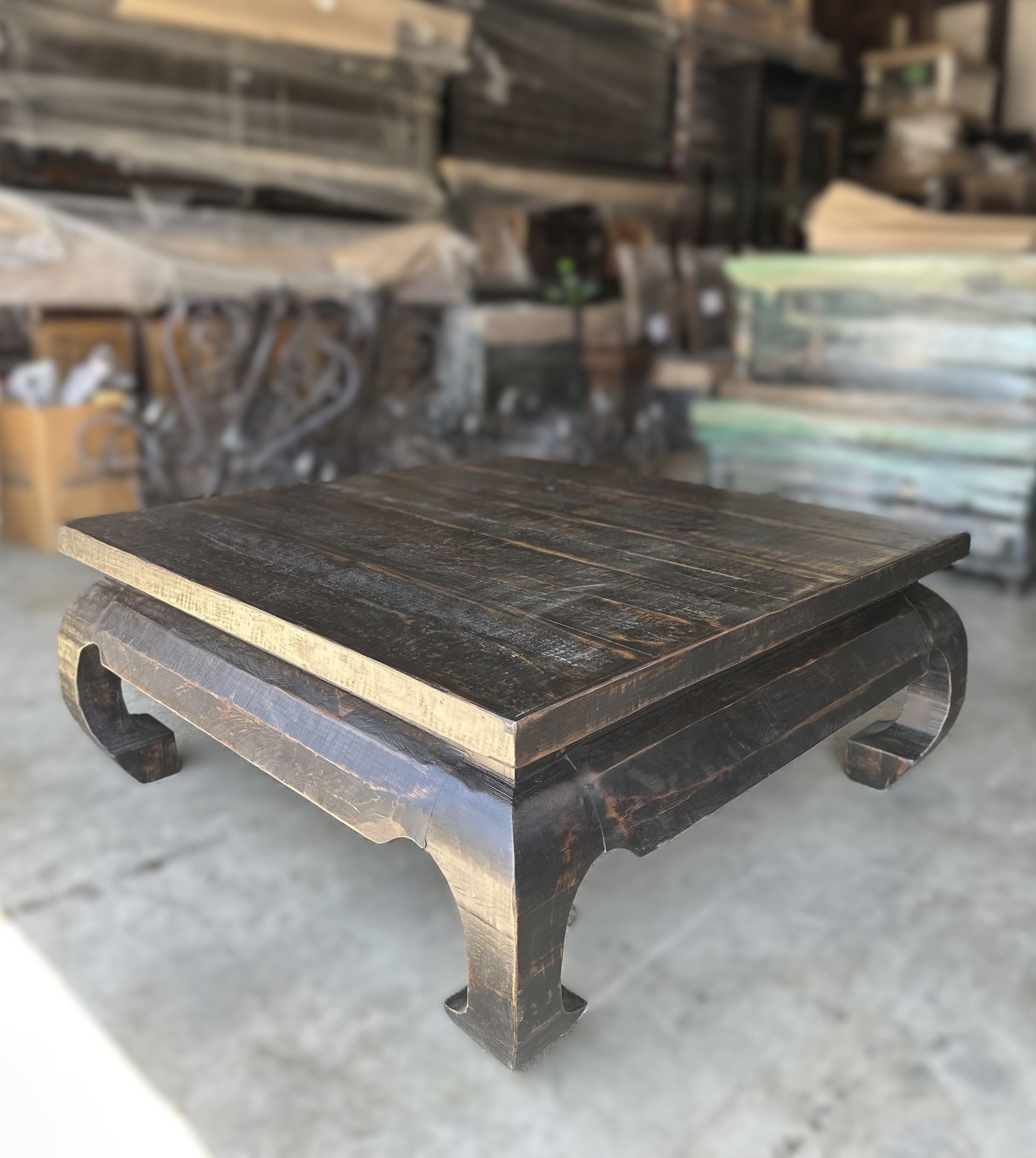 Square, dark wooden platform with curved legs, possibly a display stand, in a warehouse setting.