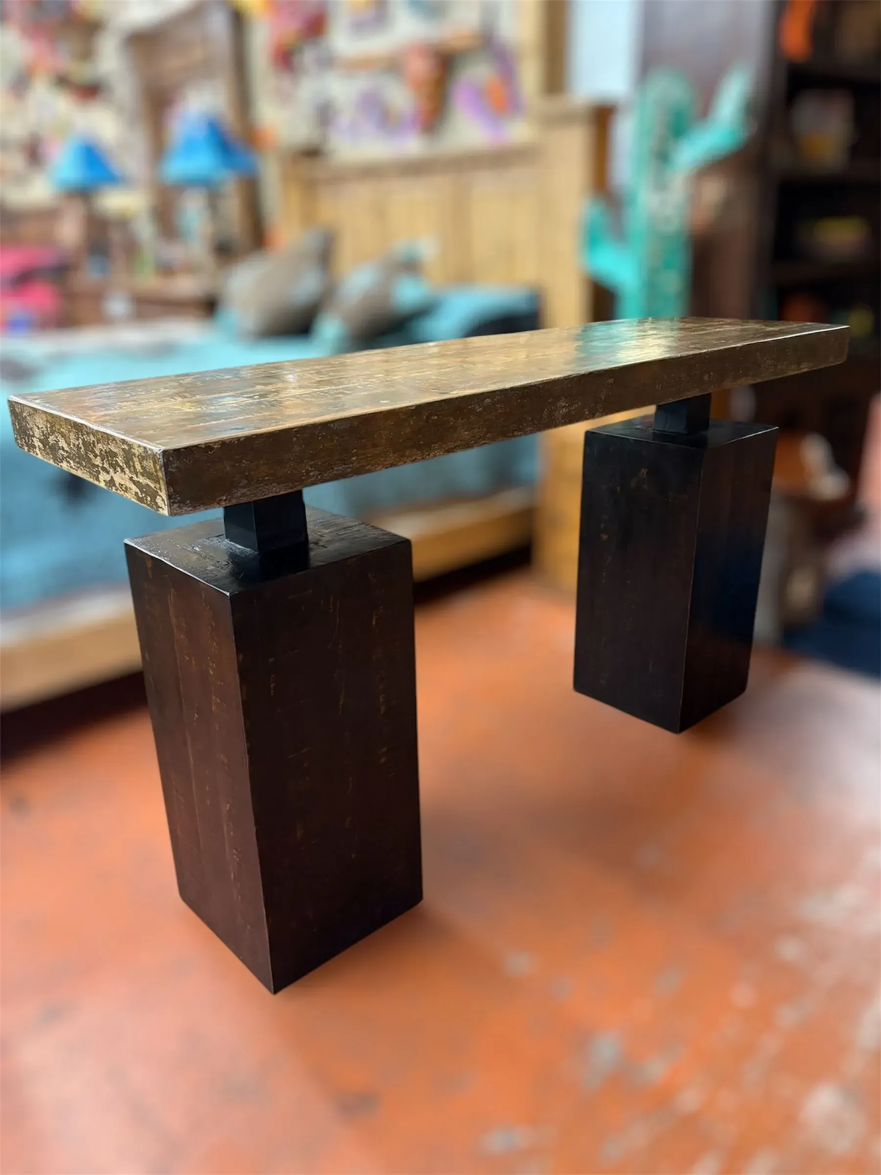 A modern, dark brown console table with a gold speckled top. The table is indoors on an orange floor, with blurred background elements.