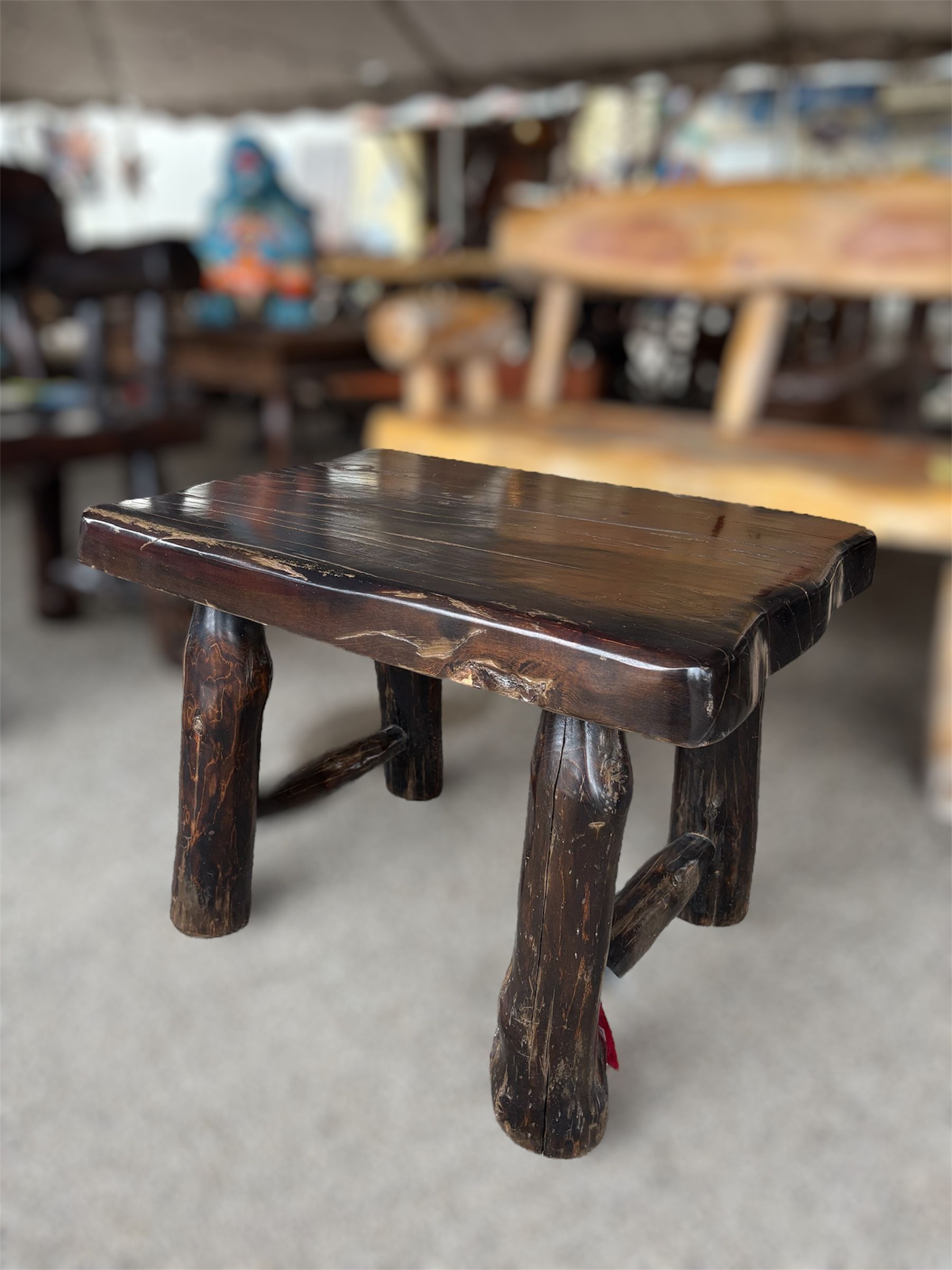 Small wooden stool with a dark, varnished top and twig legs, set in a shop with other rustic furniture.