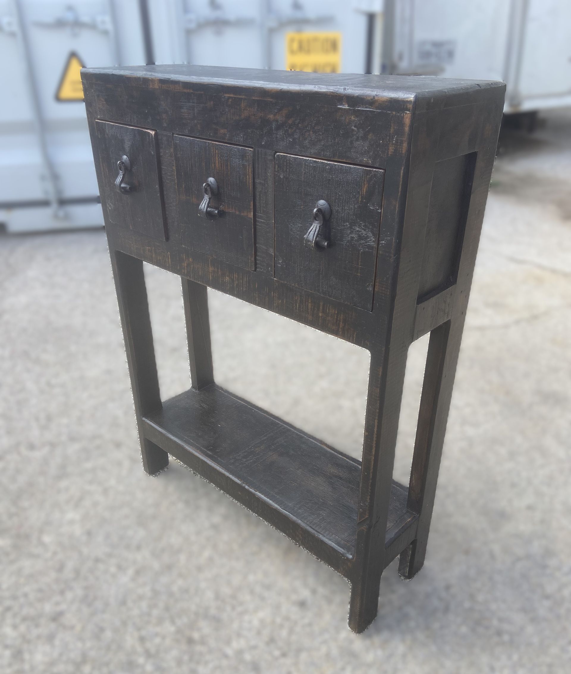Dark wooden side table with three drawers and a lower shelf. It is set outside on a concrete surface.