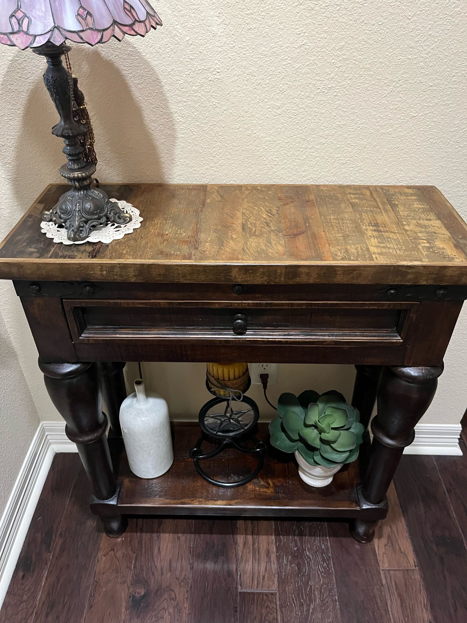 Dark wooden side table with drawer and shelf, topped with a lamp, and holding decor like a bottle and a decorative artichoke.