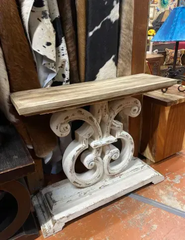 A white, ornate console table with a light wood top sits in a shop, with cowhide rugs and other furniture in the background.
