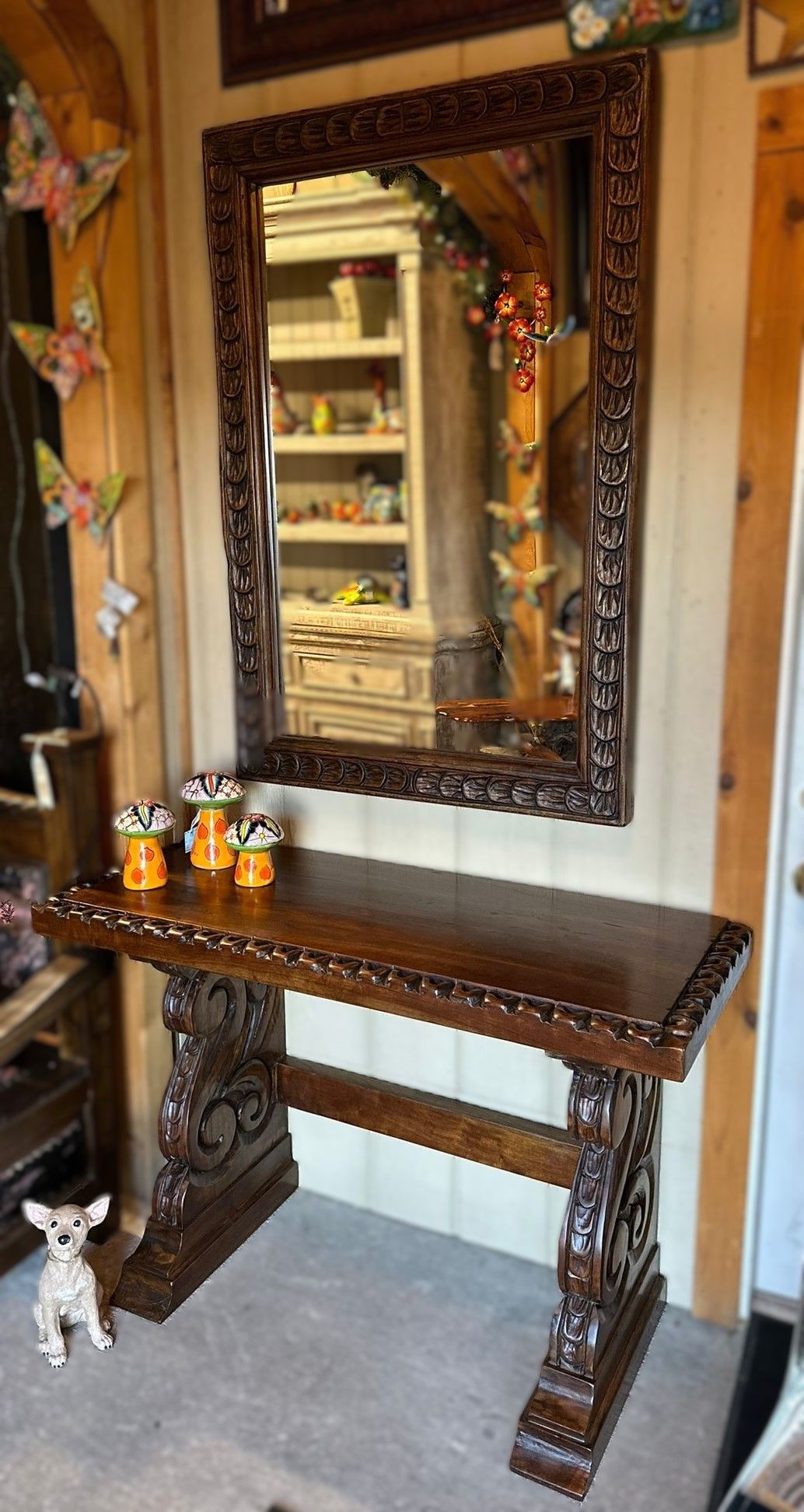Ornate wooden table and mirror. The mirror reflects shelves of items. Small statues are on the table. A small dog sits nearby.