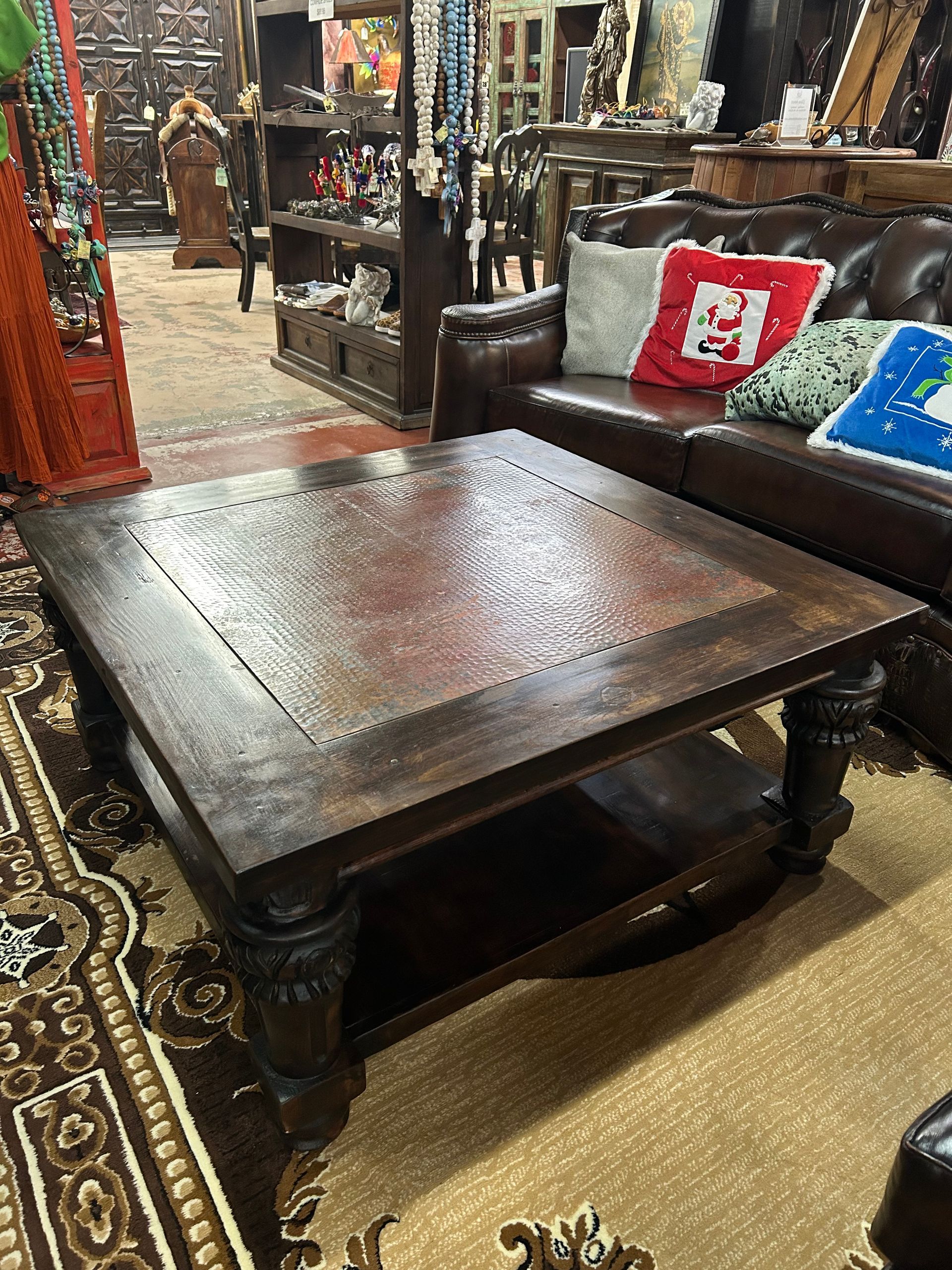 Square wooden coffee table in a shop, in front of a leather sofa. The table has a lower shelf, dark finish, and decorative legs.