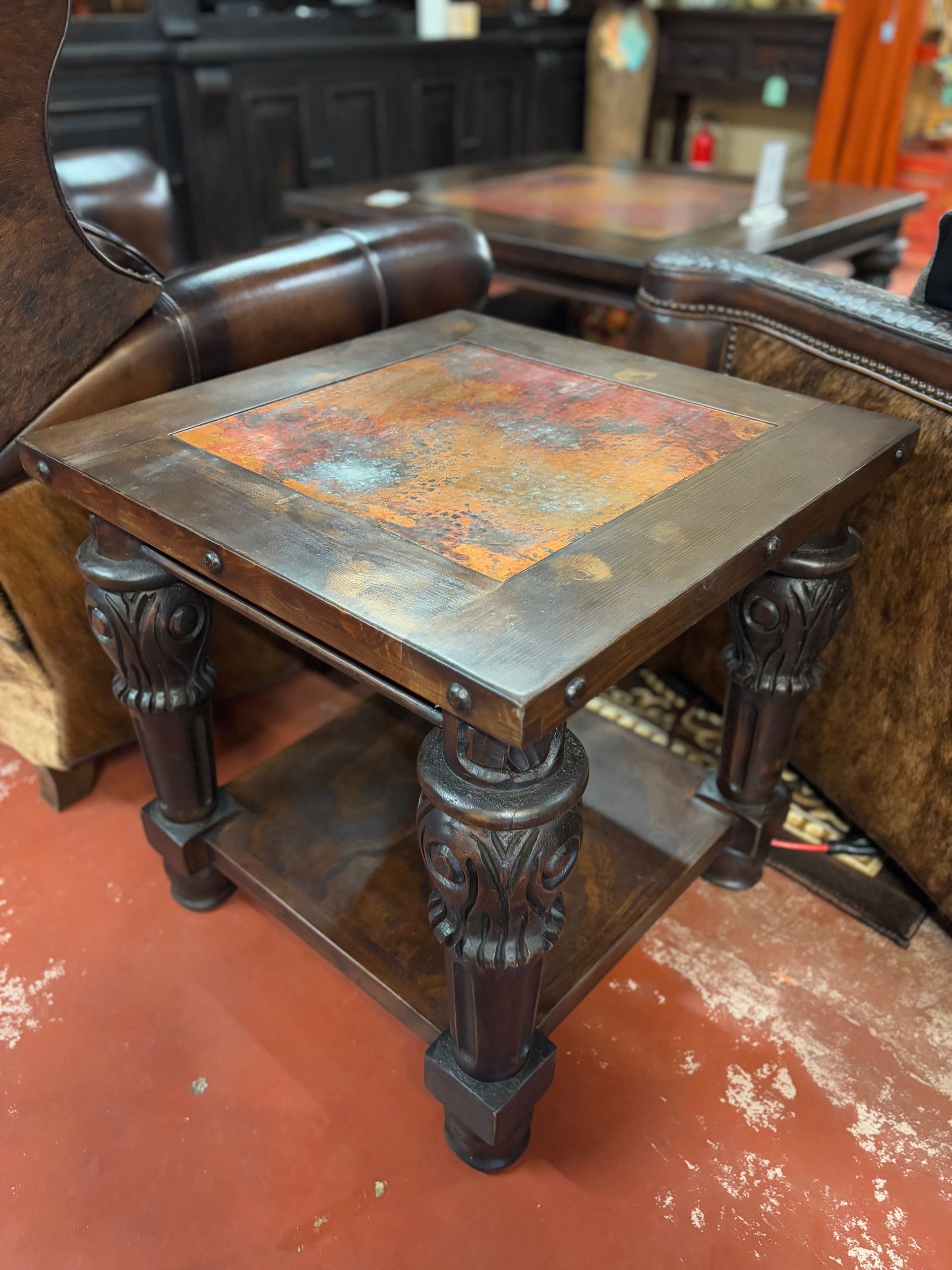 Square wooden side table with ornate legs, copper-colored top, and a lower shelf. Brown furniture is in a room.