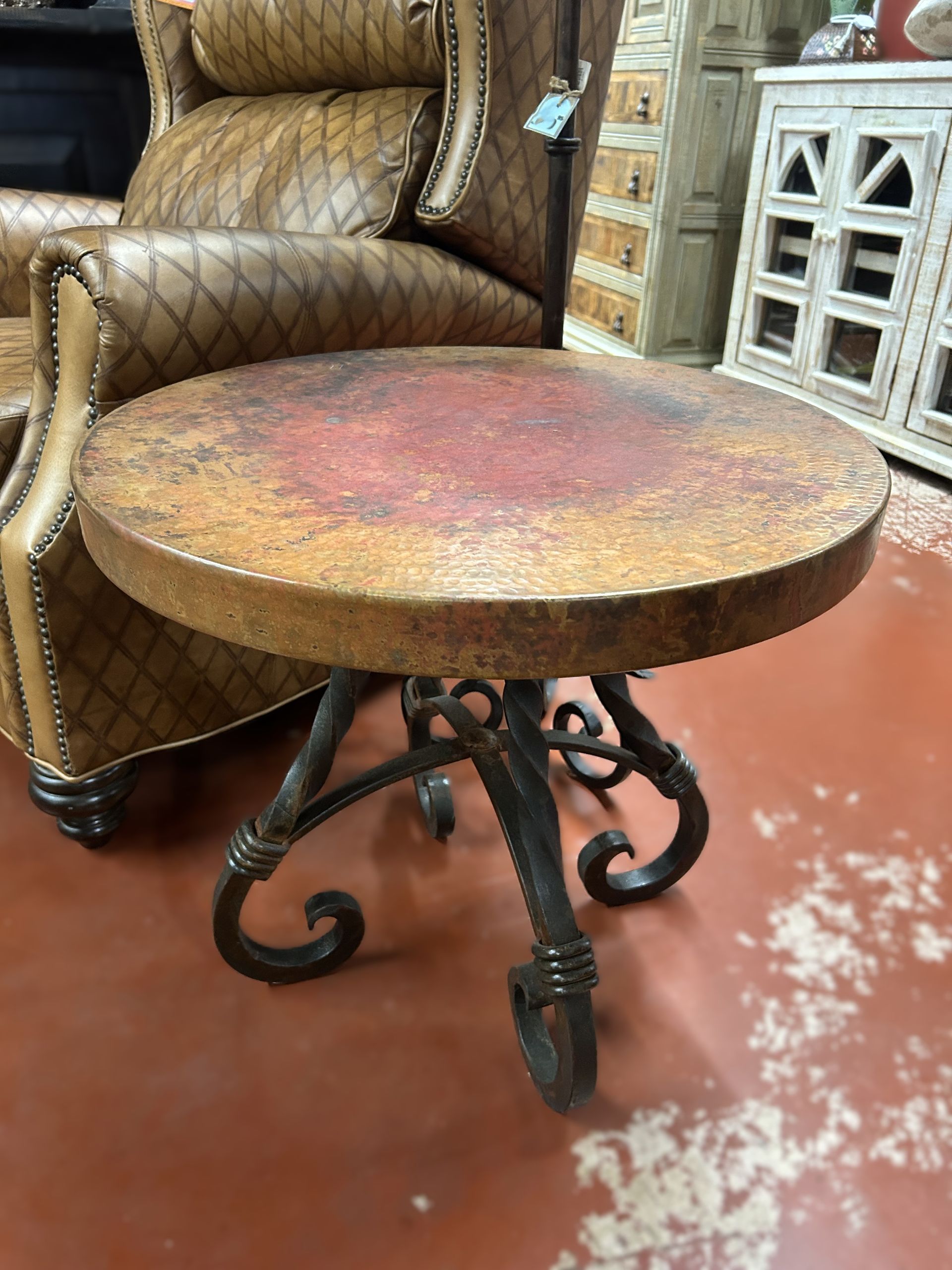 Round side table with a weathered brown and red top and wrought iron legs, beside a brown leather armchair.