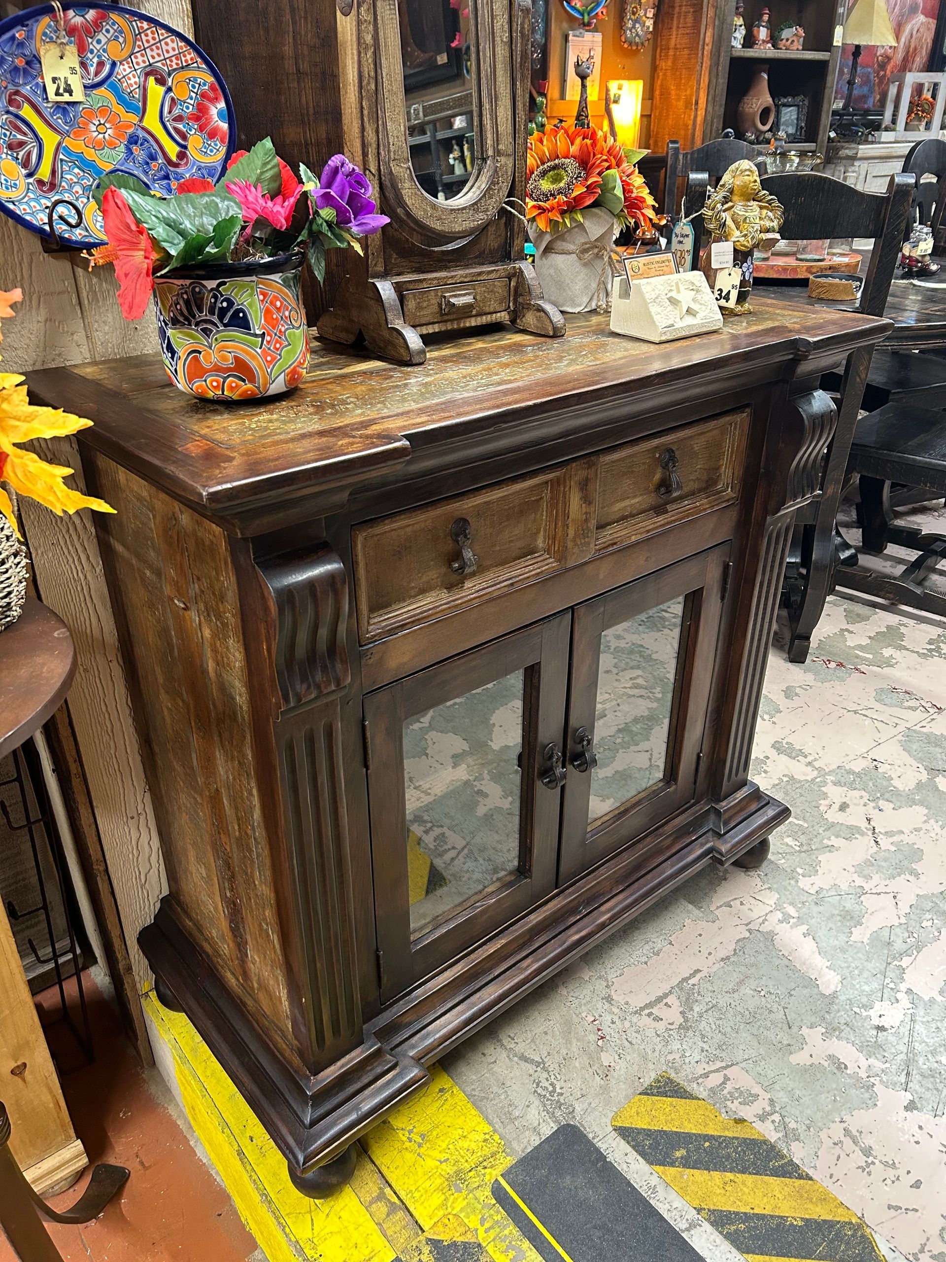 Dark wooden cabinet with mirrored doors, topped with decorative items and flowers. Set in a cluttered interior with patterned tile and warning stripes.