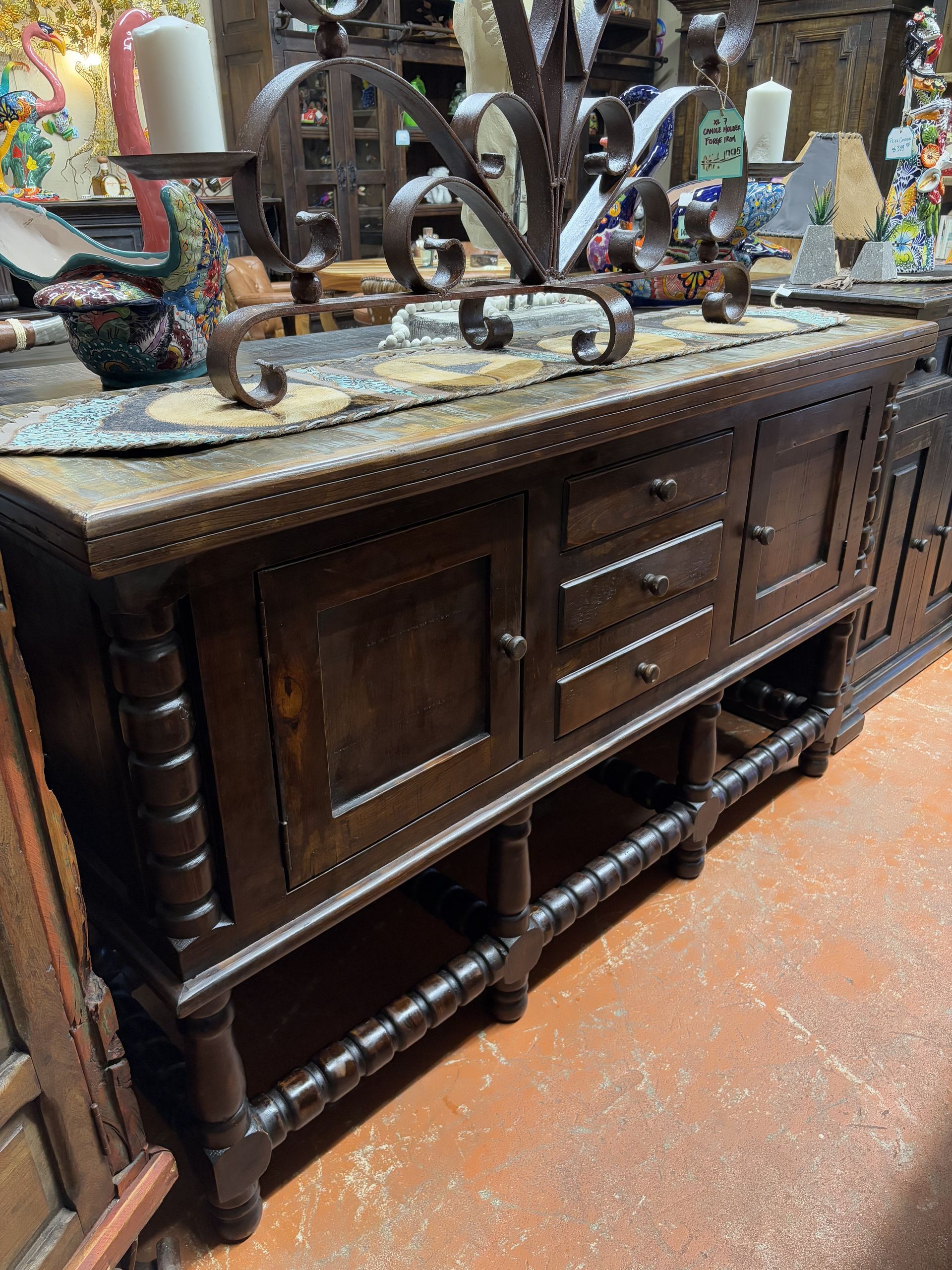 Dark wood buffet with metal accents, topped with candle holders. It has drawers and doors, sitting on a tiled floor.