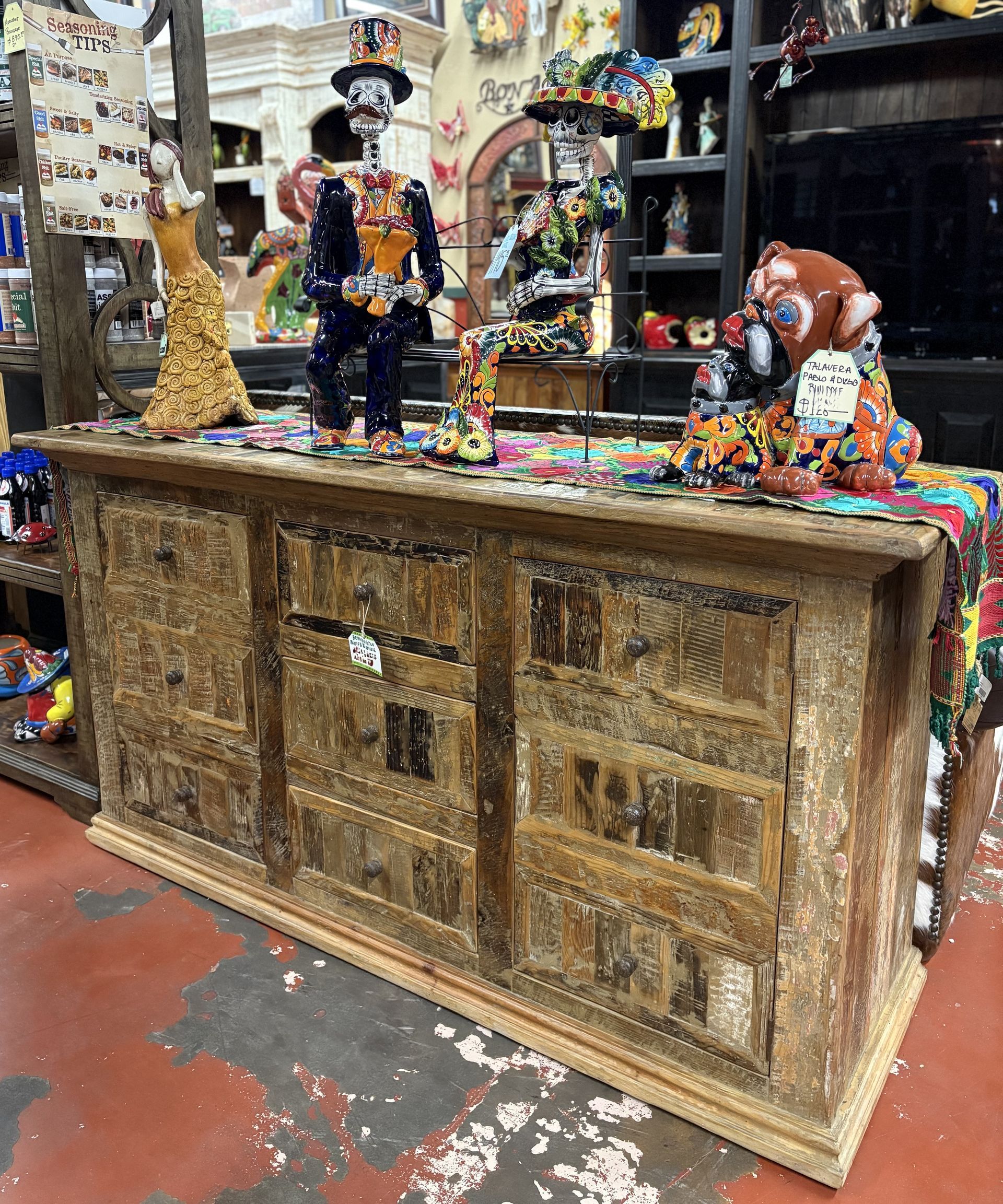 A wooden cabinet displays colorful Day of the Dead figurines and decorations in a shop. The cabinet has carved details and a worn appearance.