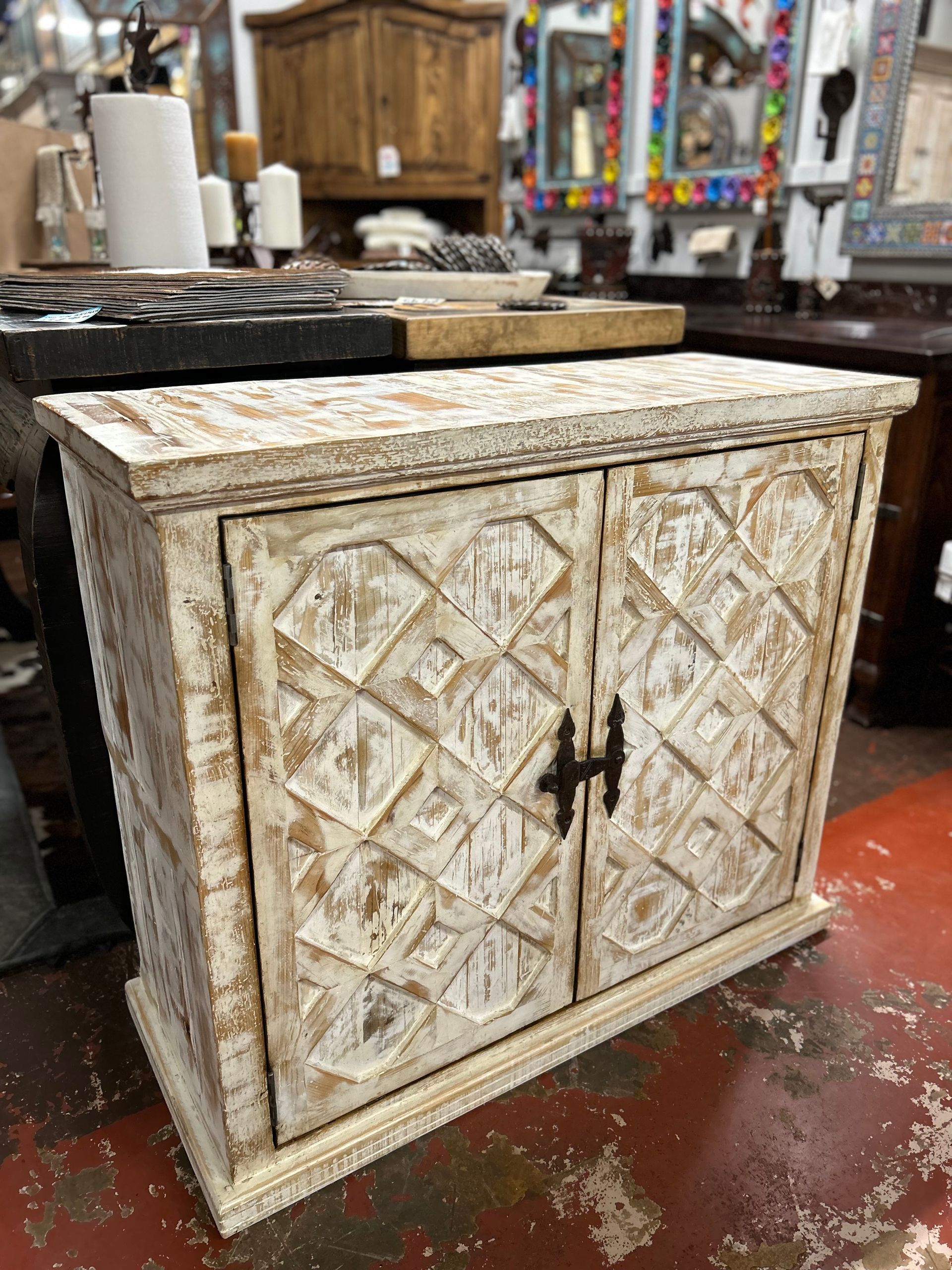 White distressed wooden cabinet with geometric carvings on the doors, black hardware, and a rustic aesthetic, displayed in a shop.