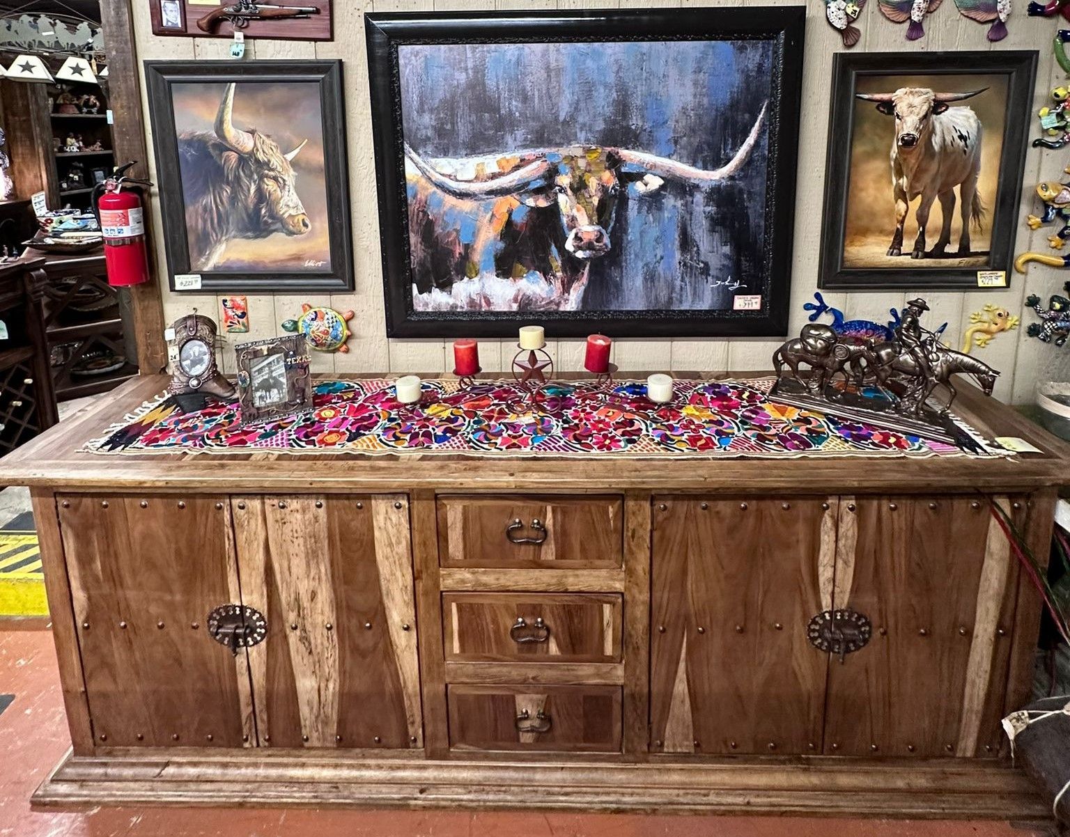 Wooden cabinet with colorful textile, candles, and art of longhorn cattle.