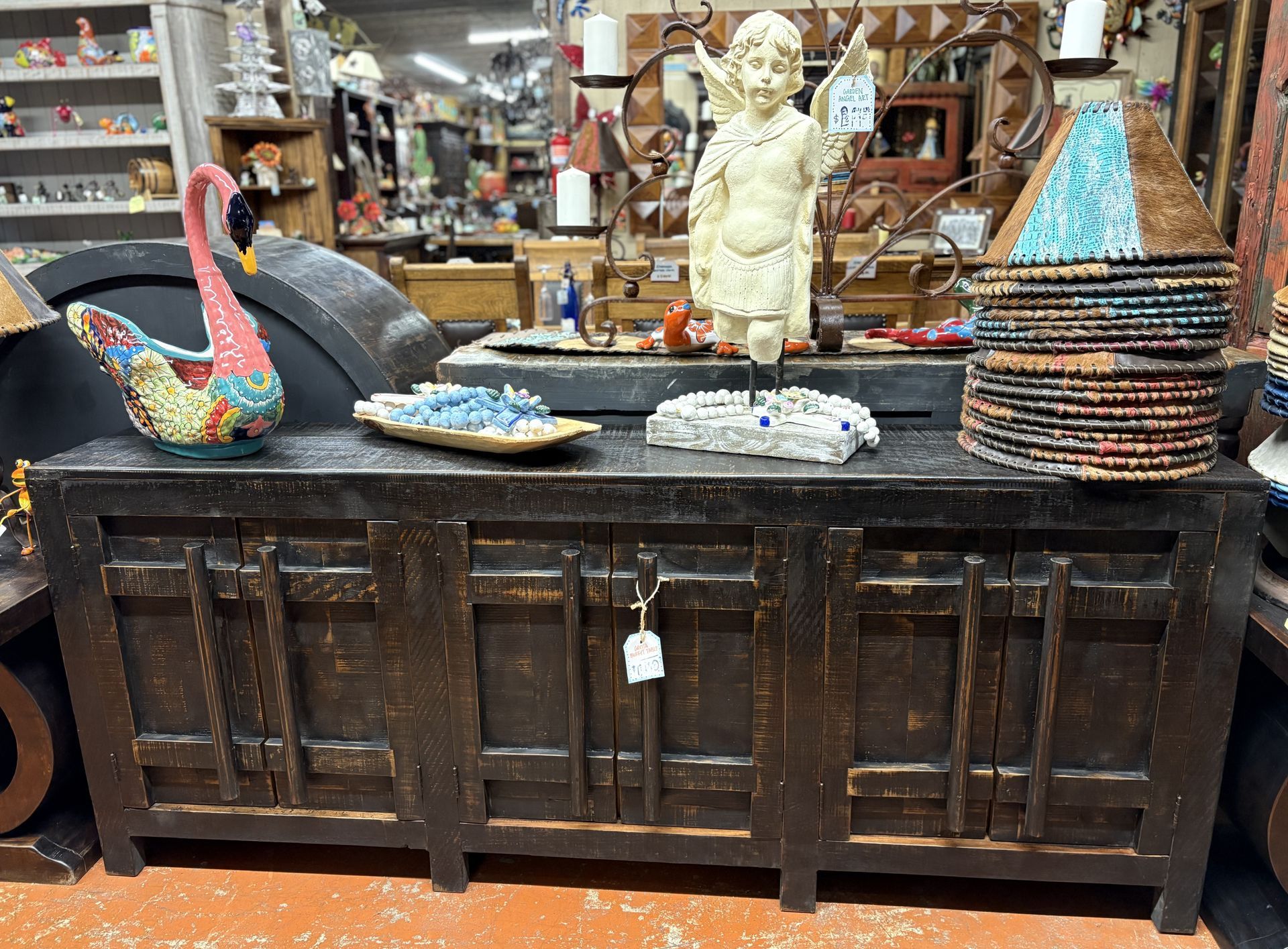 Dark wooden cabinet with decorative items on top, including a swan statue, small figurine, and stacked lampshades, in a shop setting.
