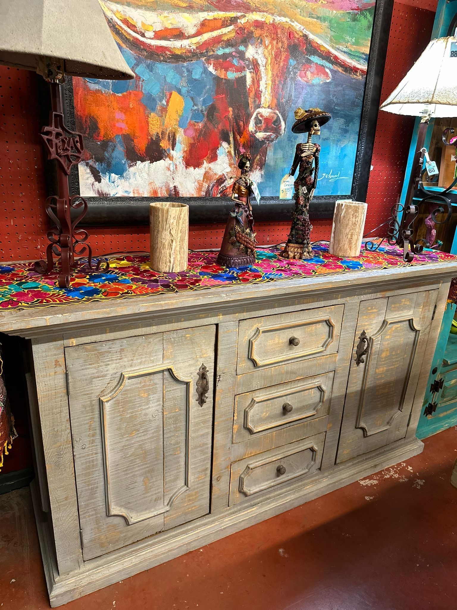 Wooden sideboard with decorative items on top, including candles and sculptures, against a colorful painting of a steer.