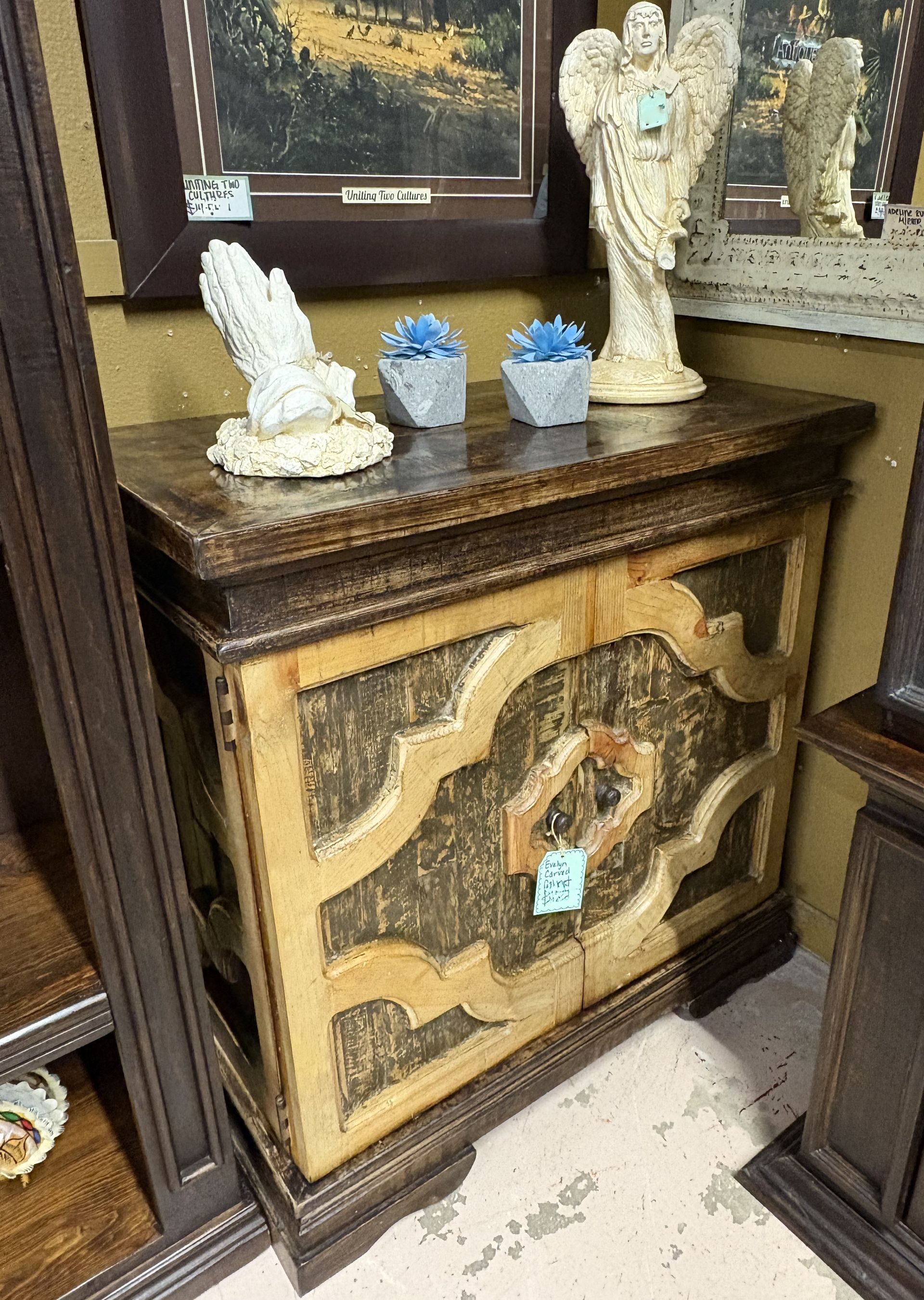 A rustic wooden cabinet with decorative front panel, topped with religious figurines and small blue succulents.