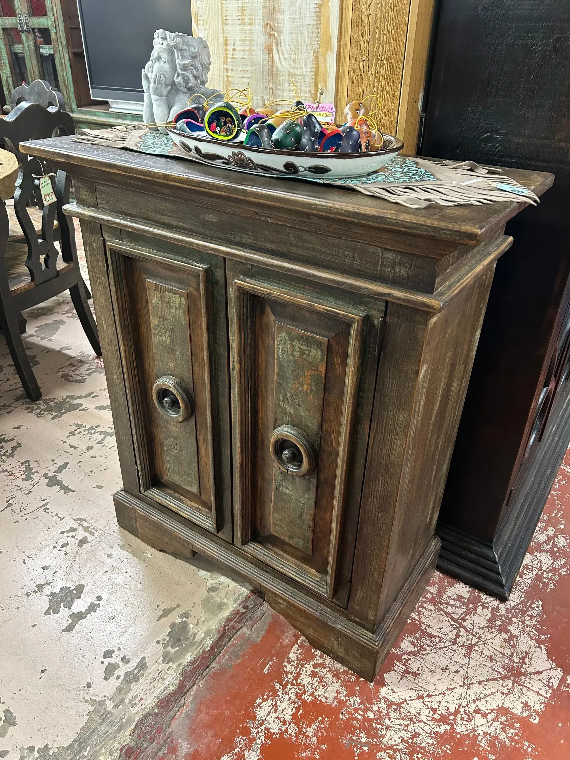 Wooden cabinet with two doors, adorned with decorative carvings and dark hardware. A bowl of colorful beads sits atop.