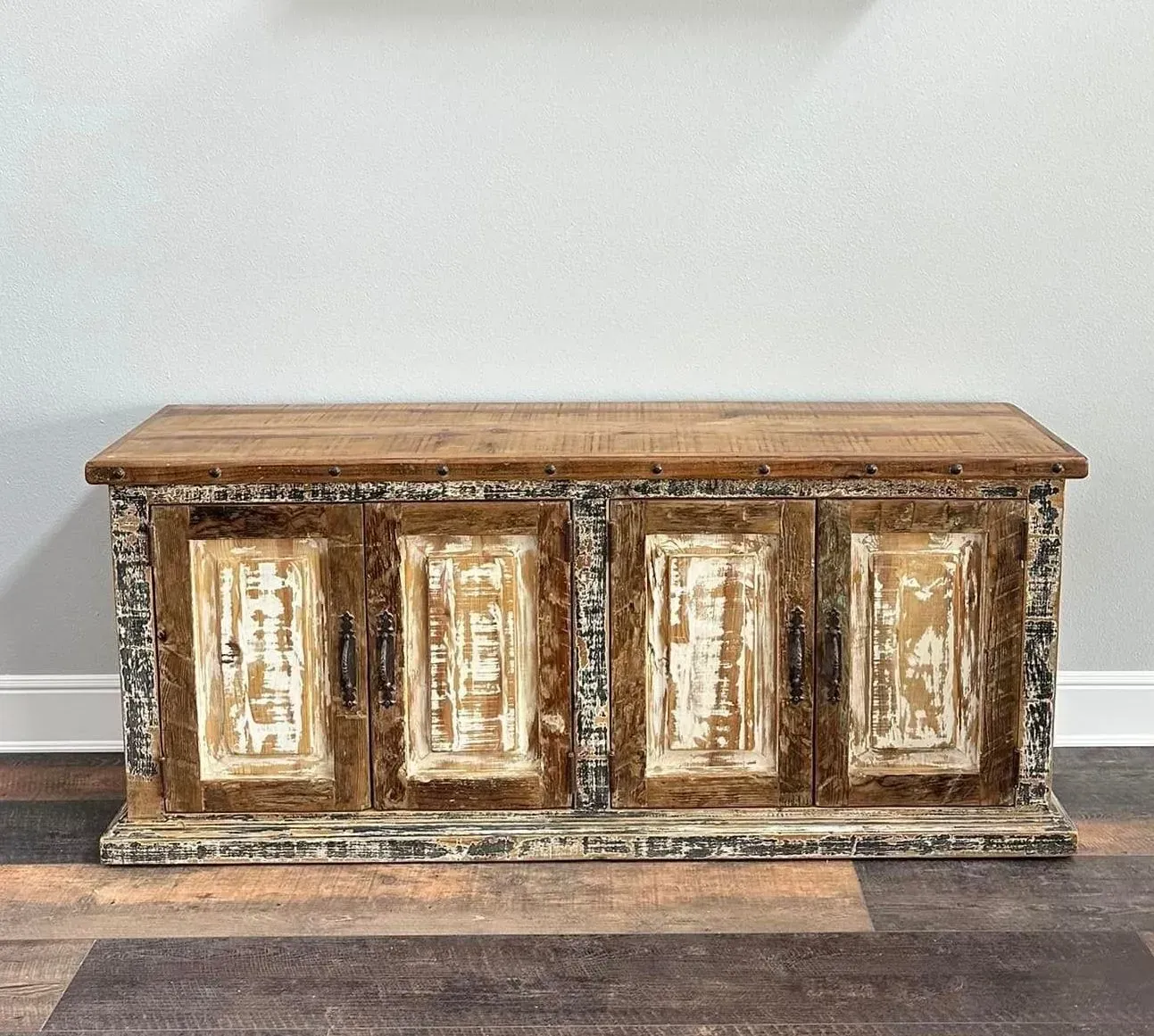 Rustic wooden cabinet with four doors, distressed white and brown paint, set against a white wall and dark floor.