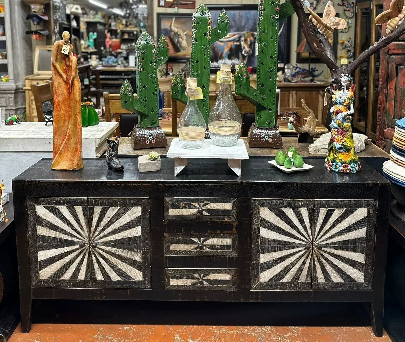 A dark wooden cabinet with sunburst patterned doors and drawers, topped with decorative items like cacti, bottles, and sculptures in a shop setting.