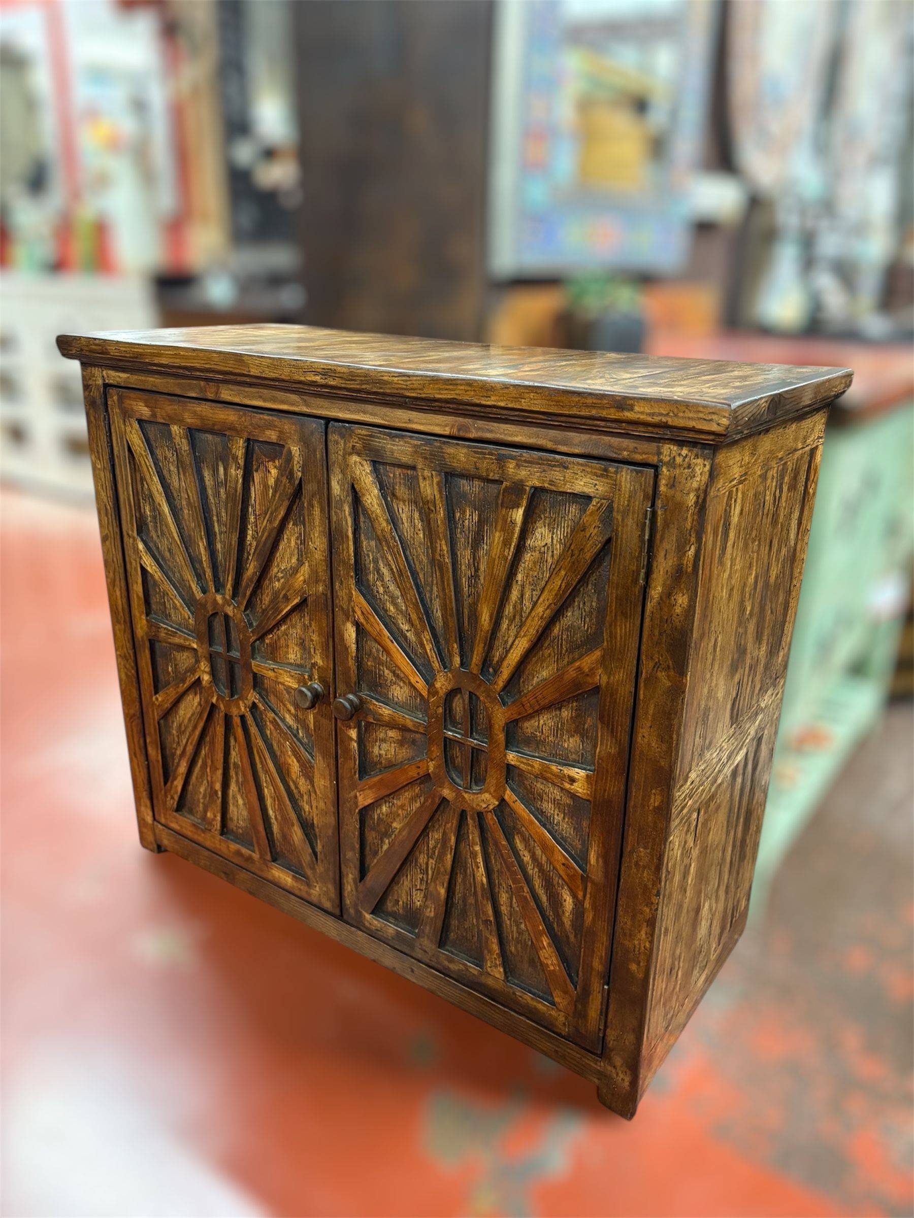 Wooden cabinet with sunburst design carved into the doors, brown with dark edges, sitting on a reddish floor, other store items in the blurry background.