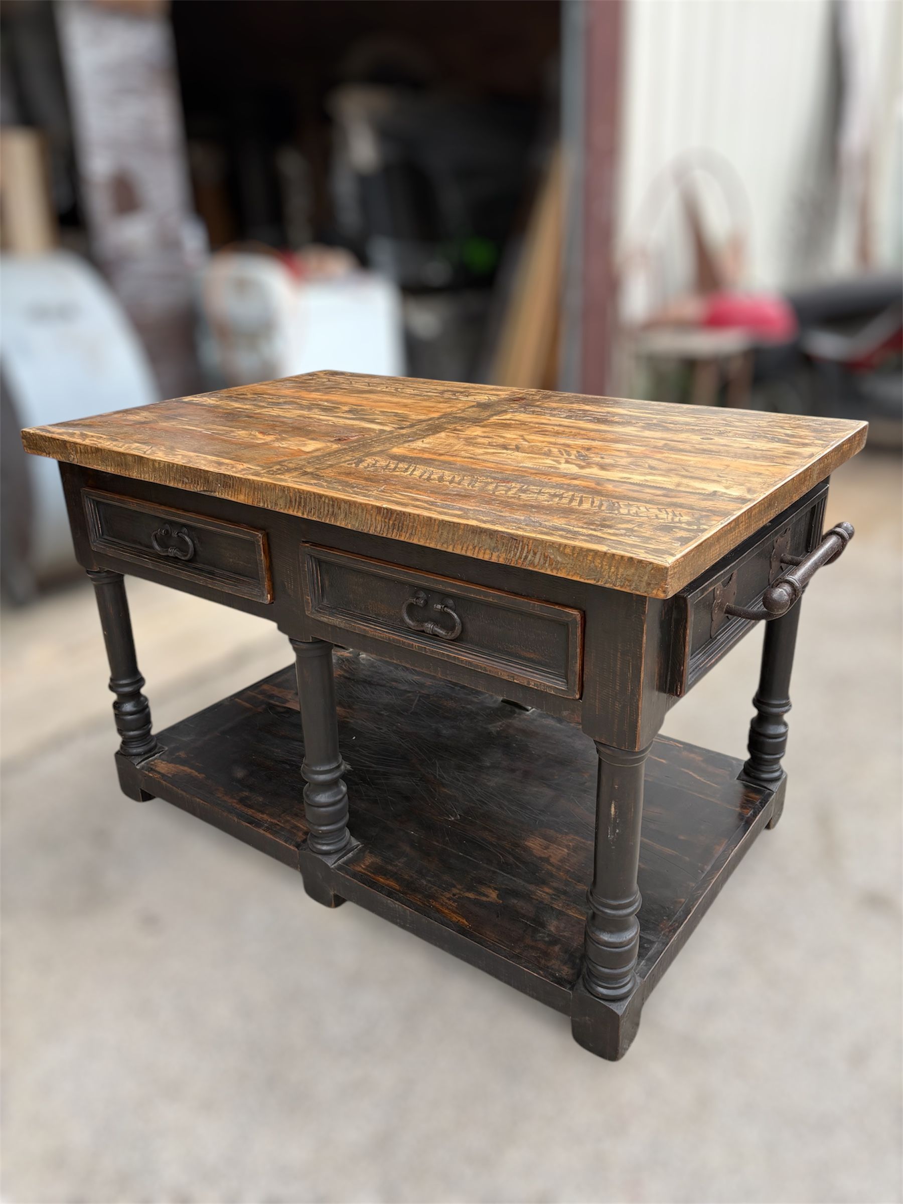 Antique kitchen island with a dark stained frame, wooden countertop, two drawers, and a lower shelf, set against a blurred workshop background.