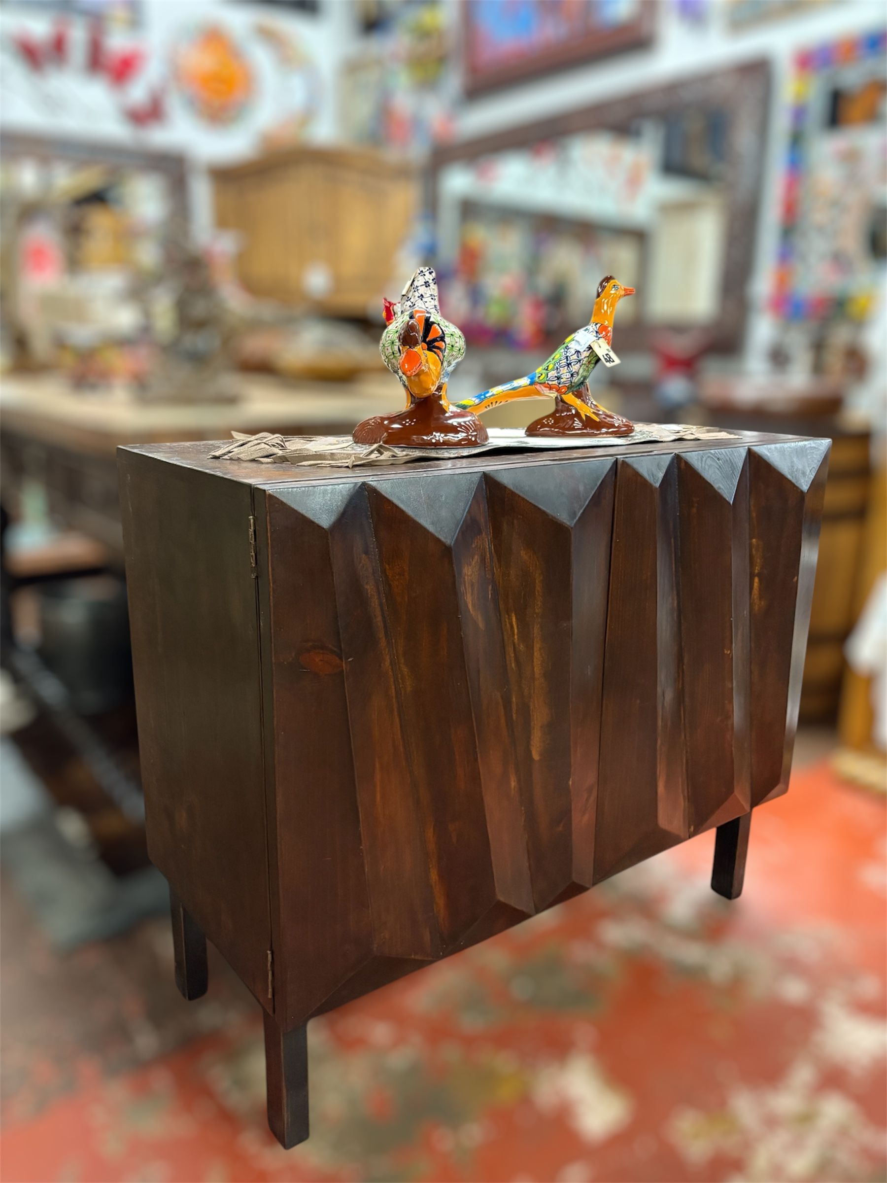 Dark wooden cabinet with zigzag top, two bird figurines, and red floor in a cluttered shop.