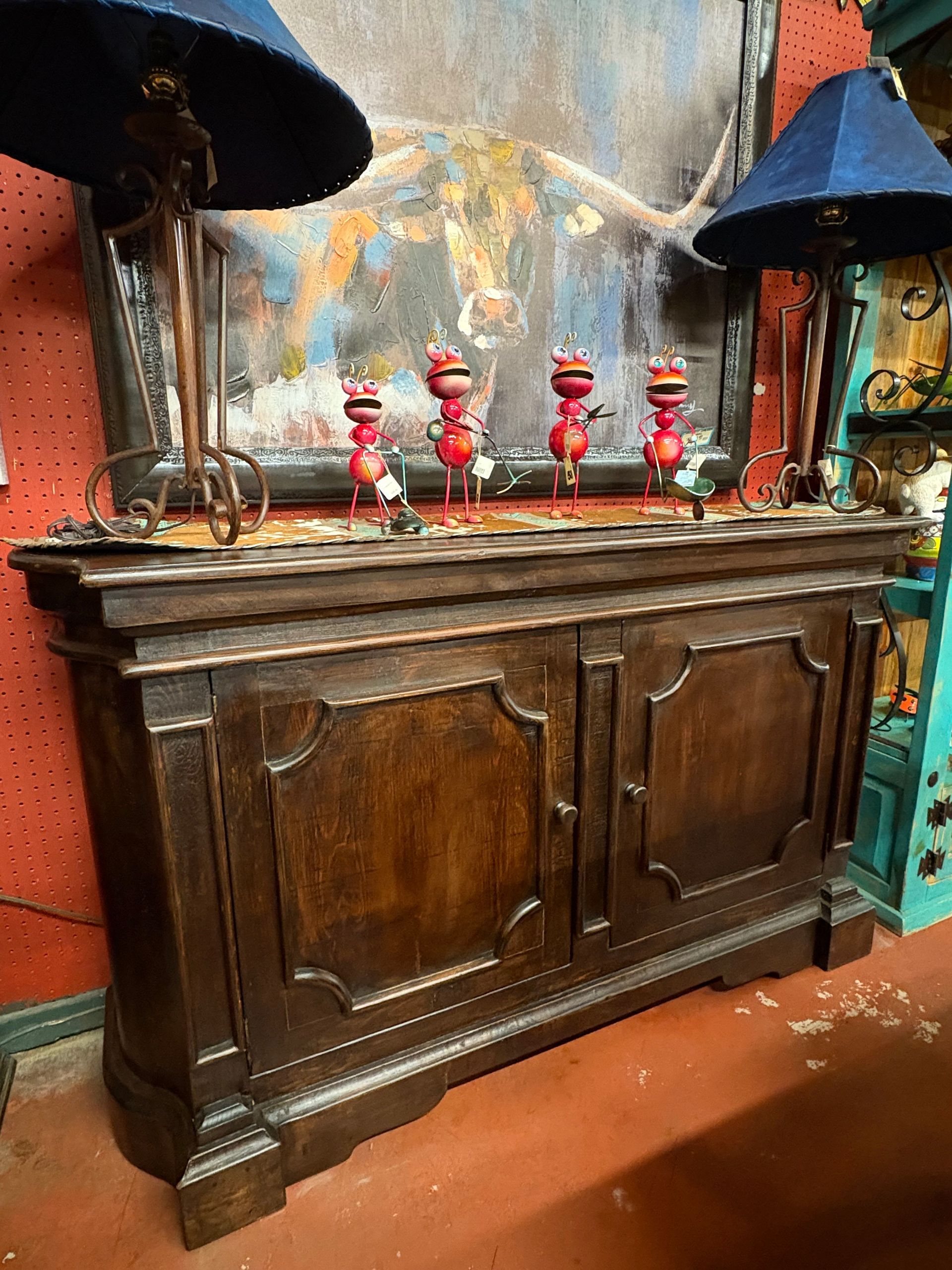 Dark wooden cabinet with a decorative front, topped with lamps and whimsical red animal figurines against a backdrop of an abstract painting.