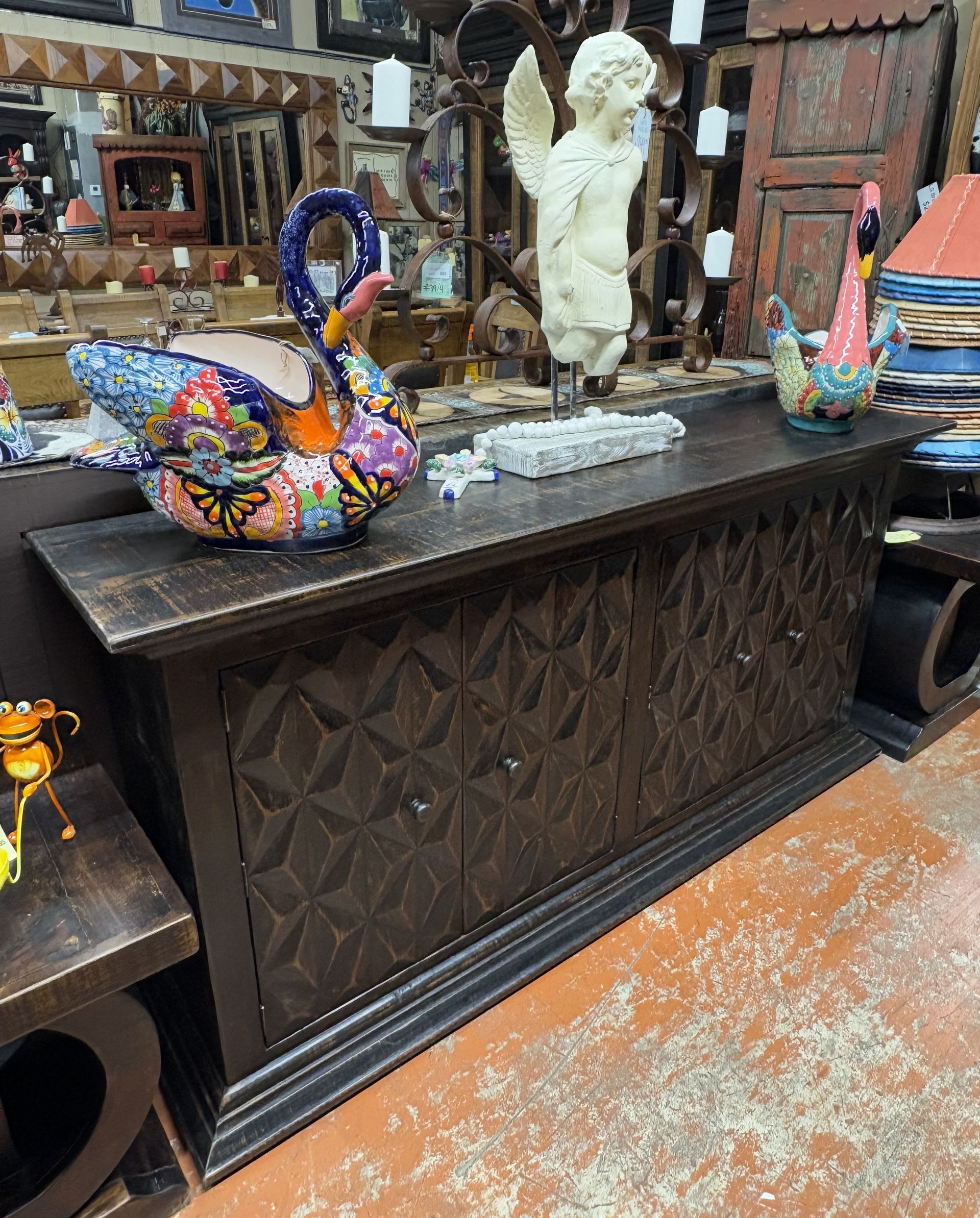 Dark wood cabinet with geometric carvings, decorated with colorful swan-shaped planter, angel statue, and other decorative items.