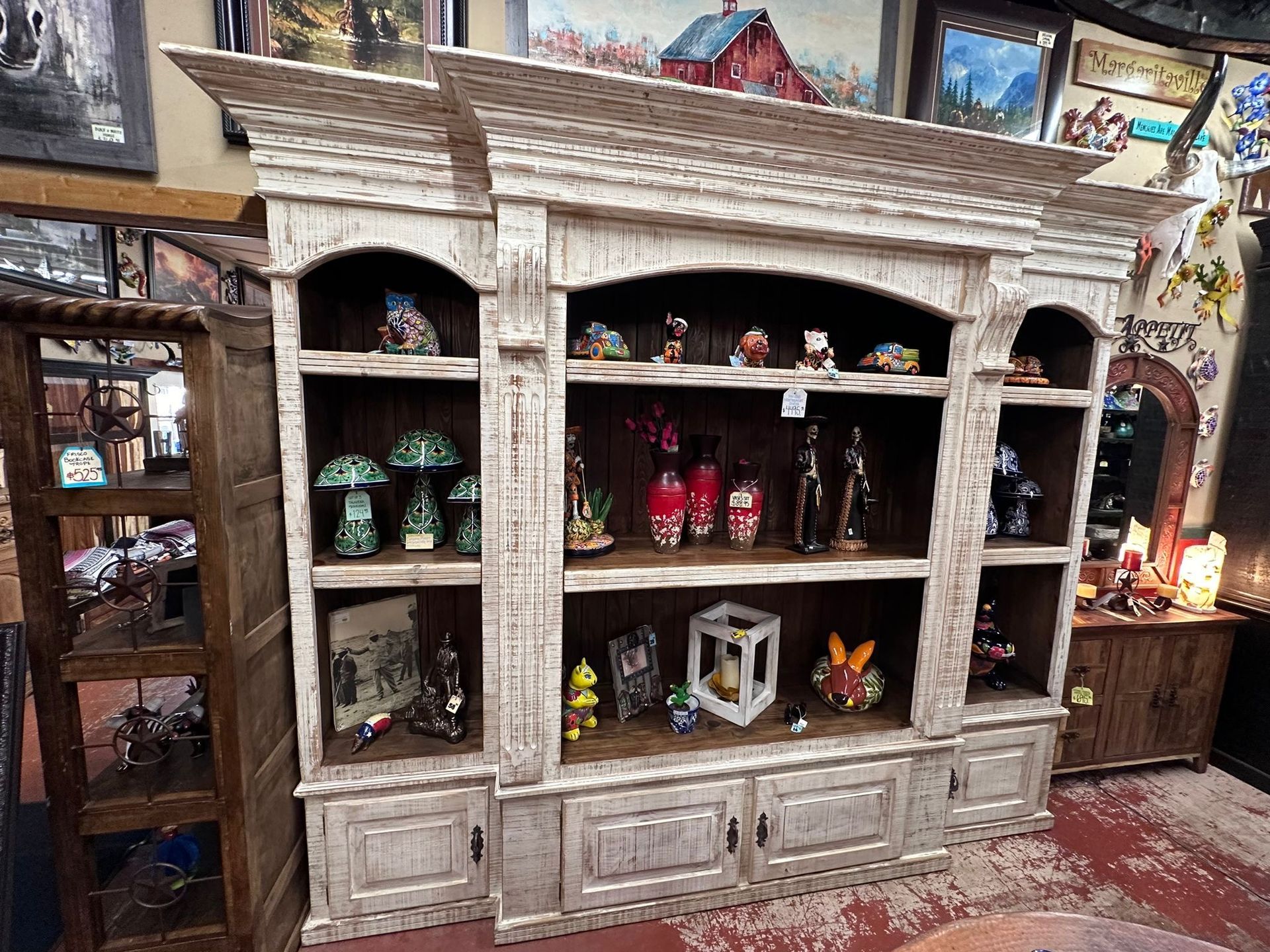 White distressed wood entertainment center with shelves and cabinets, displaying various decorative items in a store setting.