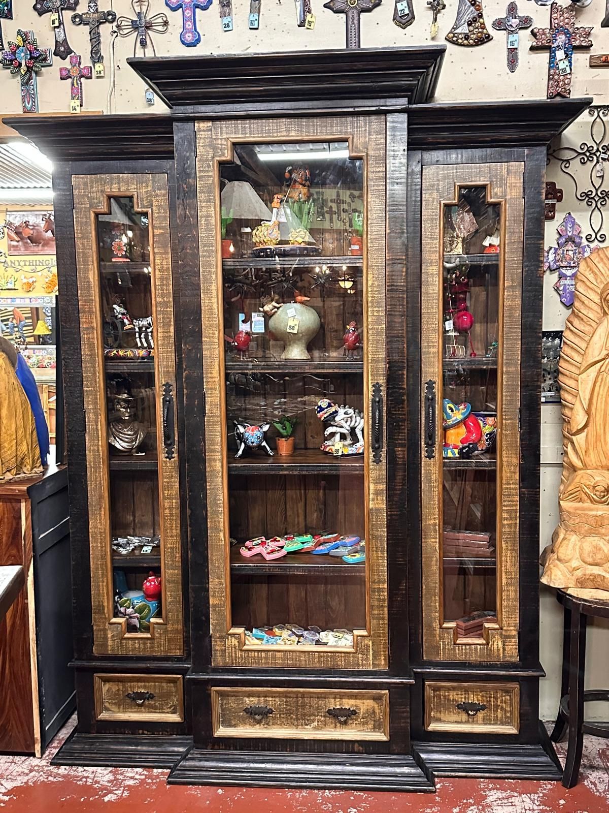 Tall, dark wood cabinet with glass doors displaying various figurines. The cabinet is in a shop setting, surrounded by other items.