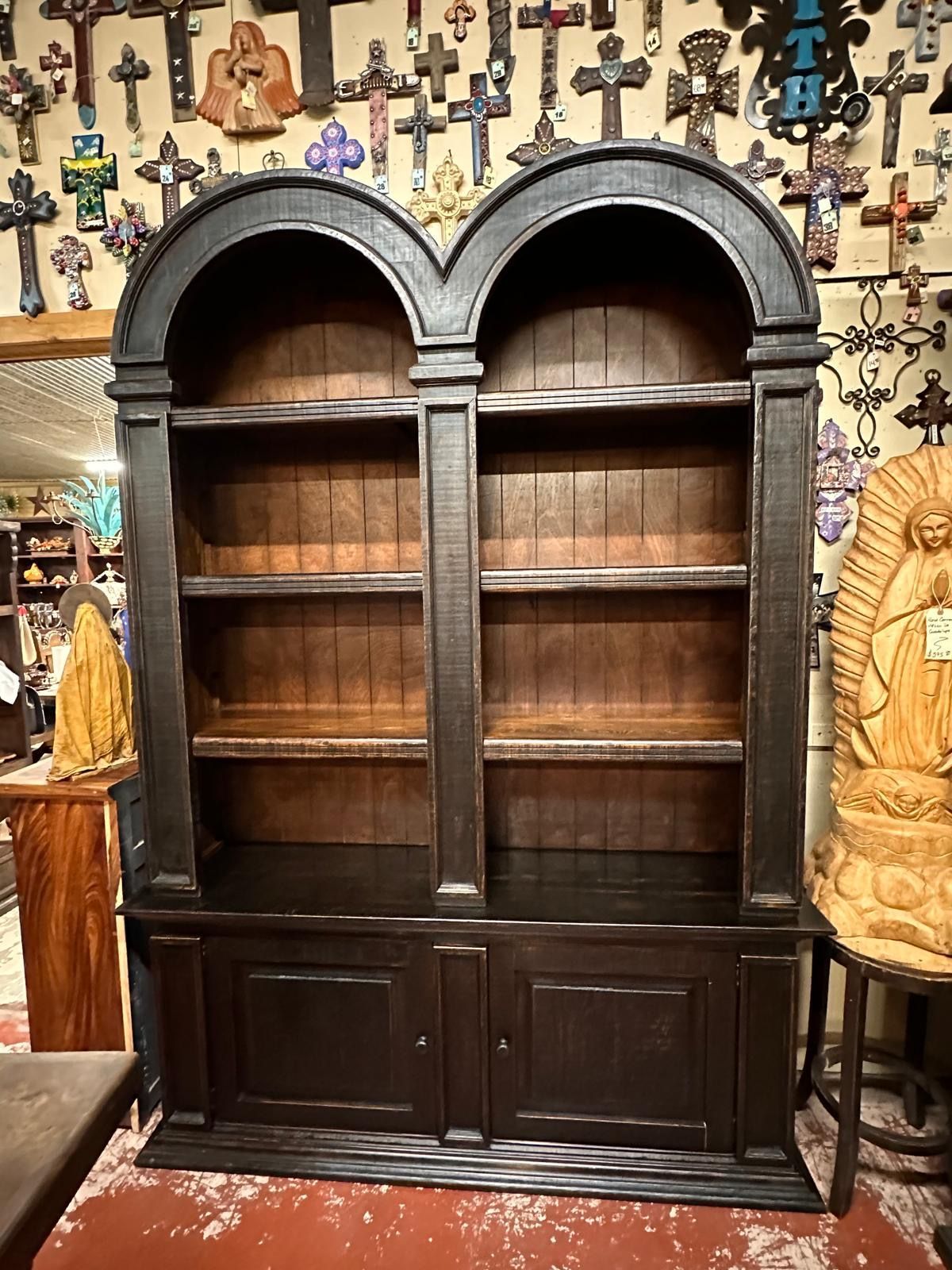 Black wooden bookshelf with arched top and four shelves, flanked by two cabinet doors. The background has many crosses.