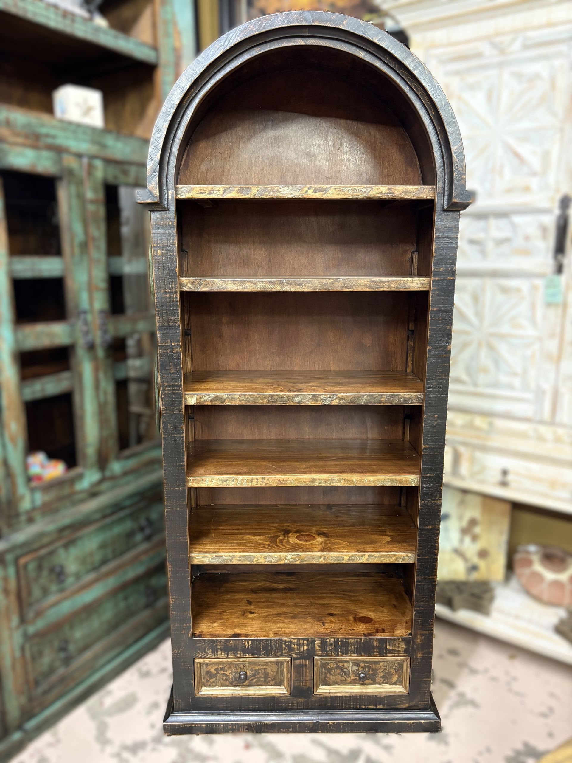 Tall, arched wooden bookshelf with six shelves and two drawers, painted black and gold. It stands in a shop setting with other furniture visible.