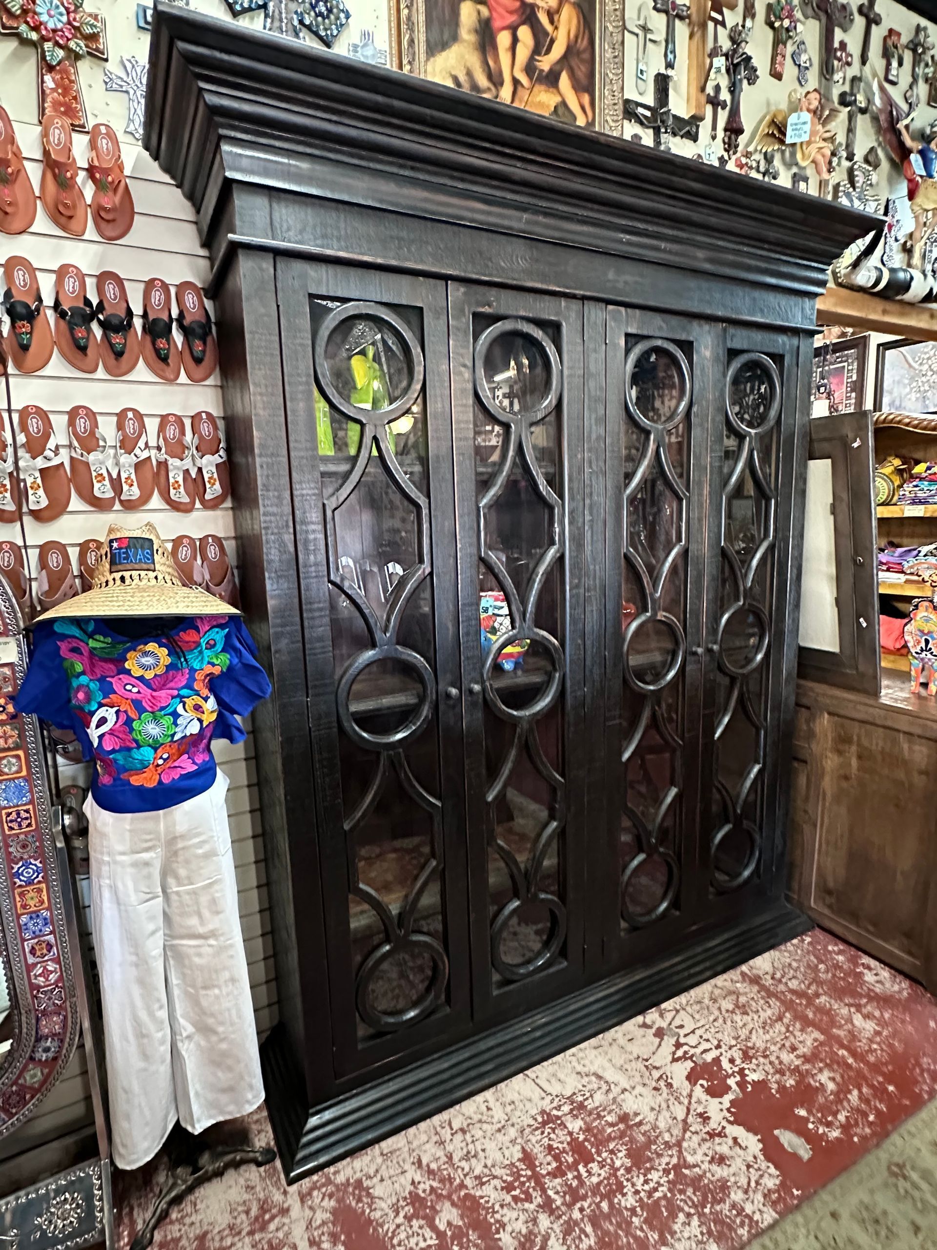 Dark wooden cabinet with glass doors and geometric design; clothing and other items surround it in a store.