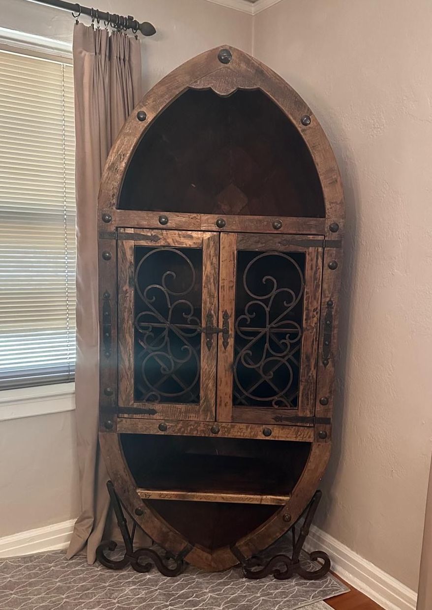 Wooden boat-shaped cabinet with ornate metal accents and glass doors in a room. The cabinet is brown, with decorative metal trim.