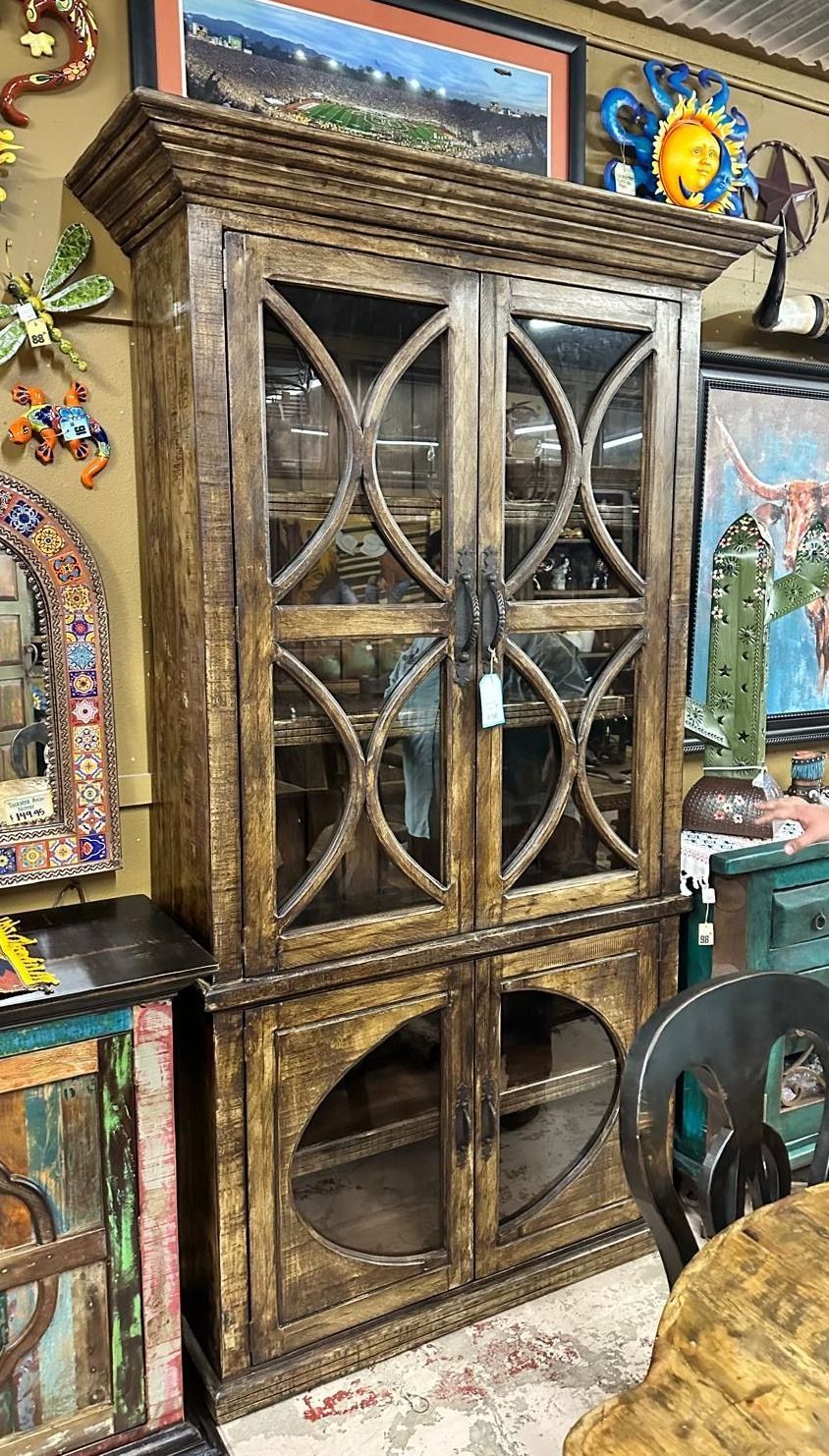 Tall, rustic wooden cabinet with glass doors and decorative circular accents, in a shop setting with other furniture and art.