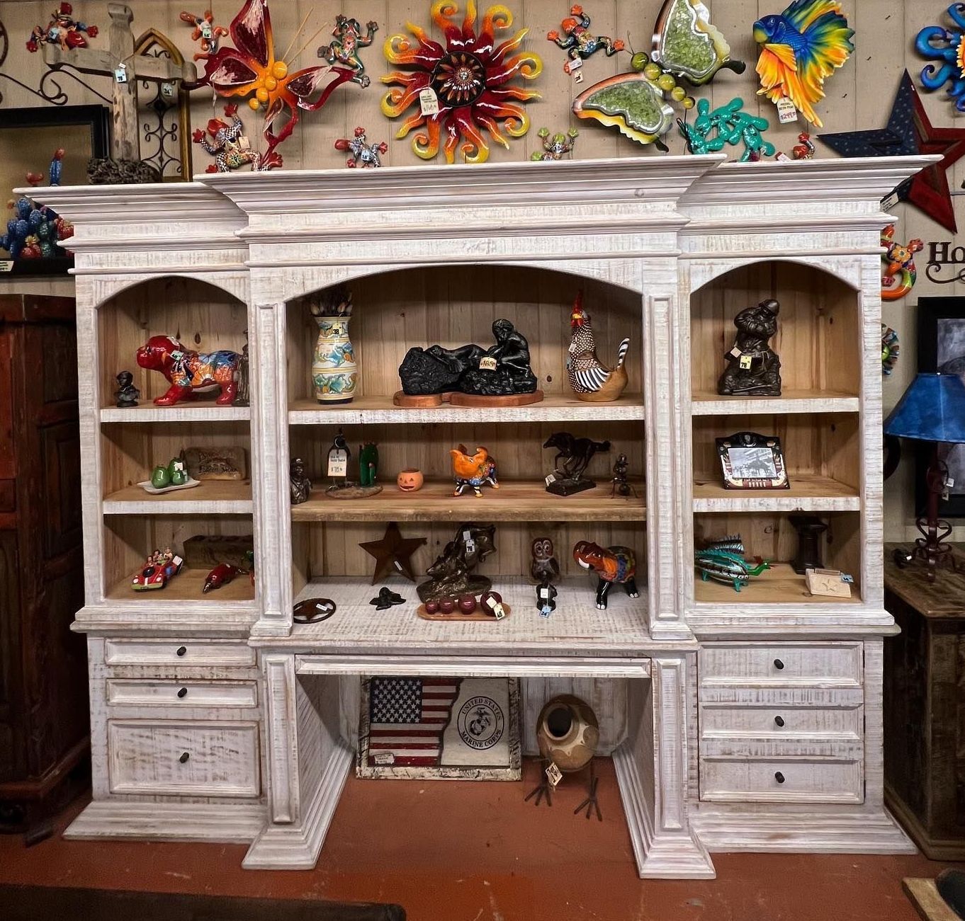 White distressed wooden desk with upper shelving, displaying various decorative items, set against a wall with colorful metal art.