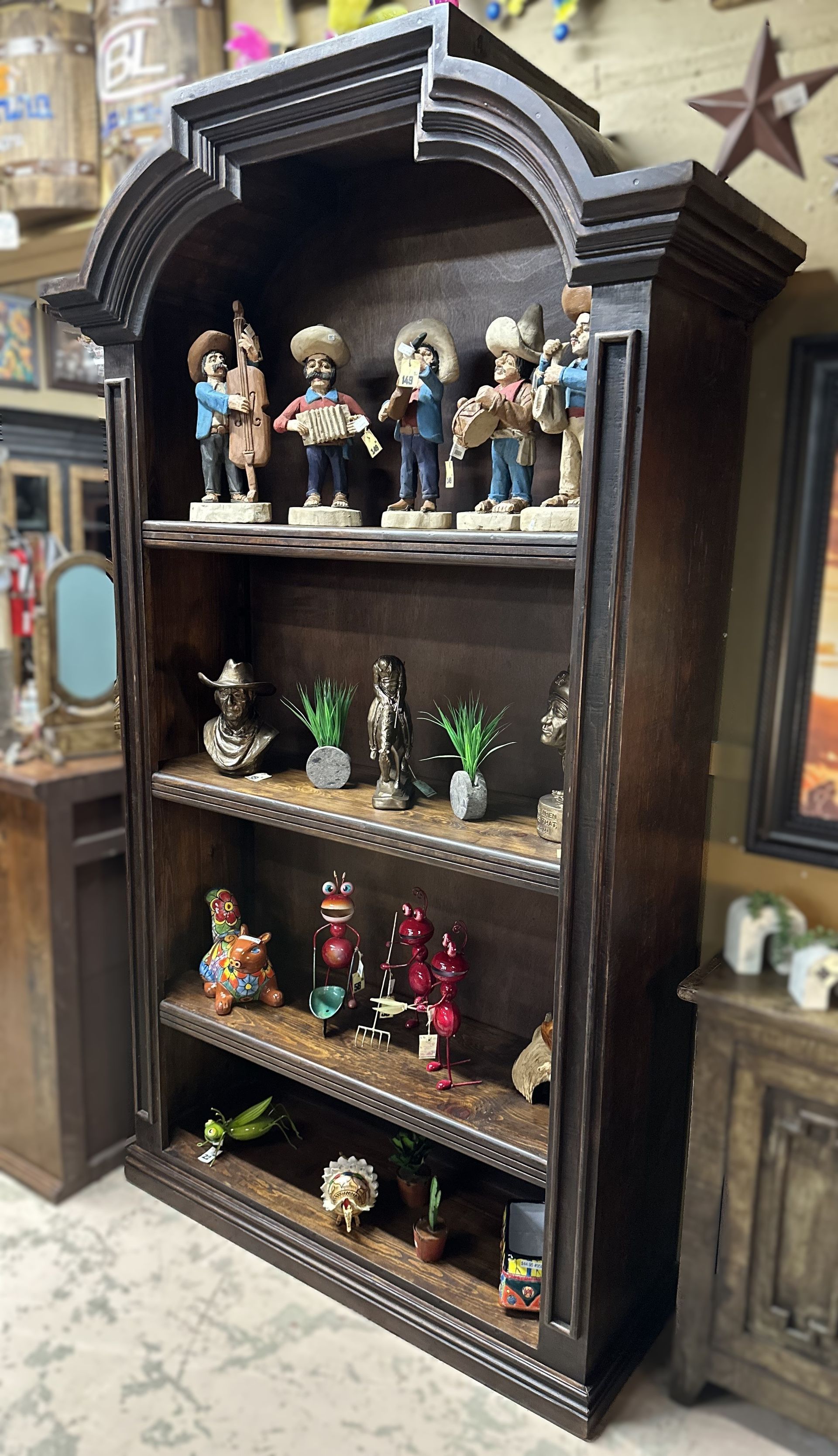 Dark wooden bookshelf filled with figurines, including skeletons and plants, in a shop setting. The shelf has an arched top.