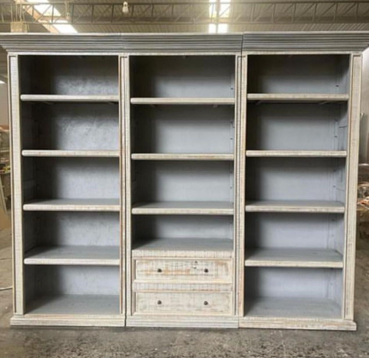 Three-section gray bookcase with open shelves and two drawers in the center section; the wood has a distressed finish.