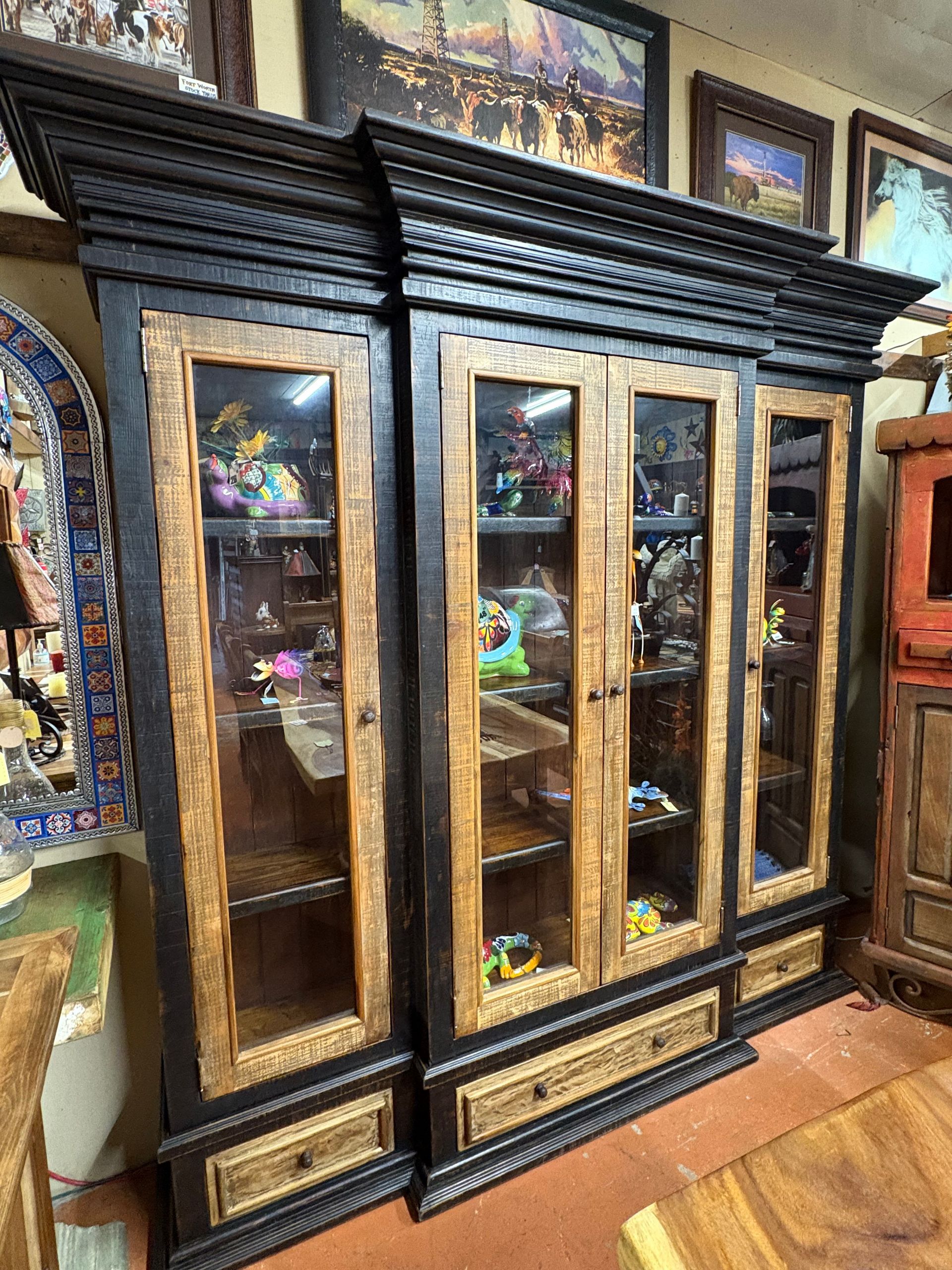 Three dark wood cabinets with glass doors and lower drawers, filled with items in a store setting.