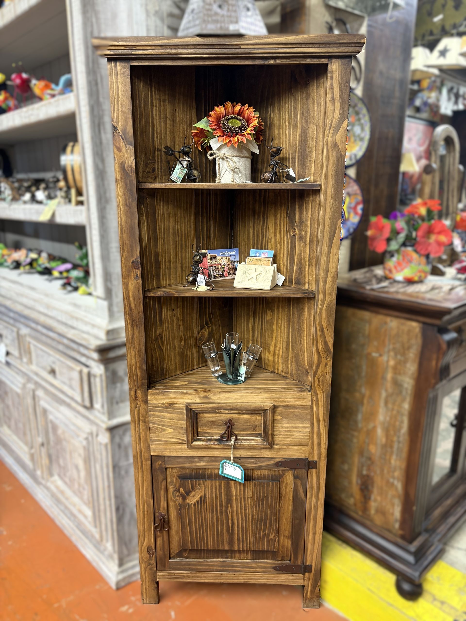 Rustic, wooden corner cabinet with shelves holding decorations; includes a drawer and lower cabinet. The cabinet is in a shop setting.