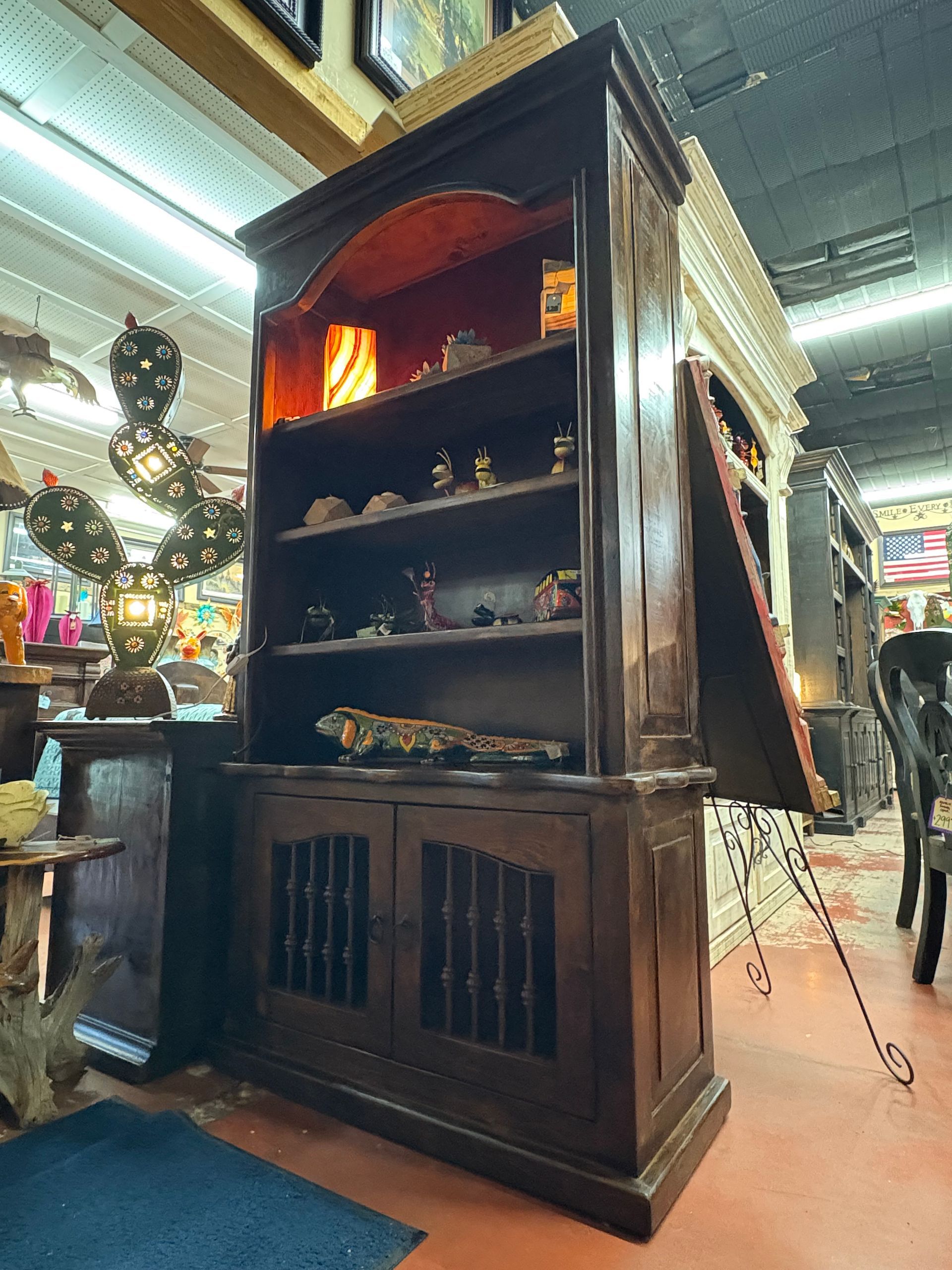 Dark wood bookcase with open shelves displaying items. The cabinet has a hinged door that is open to the side. The setting appears to be a store.