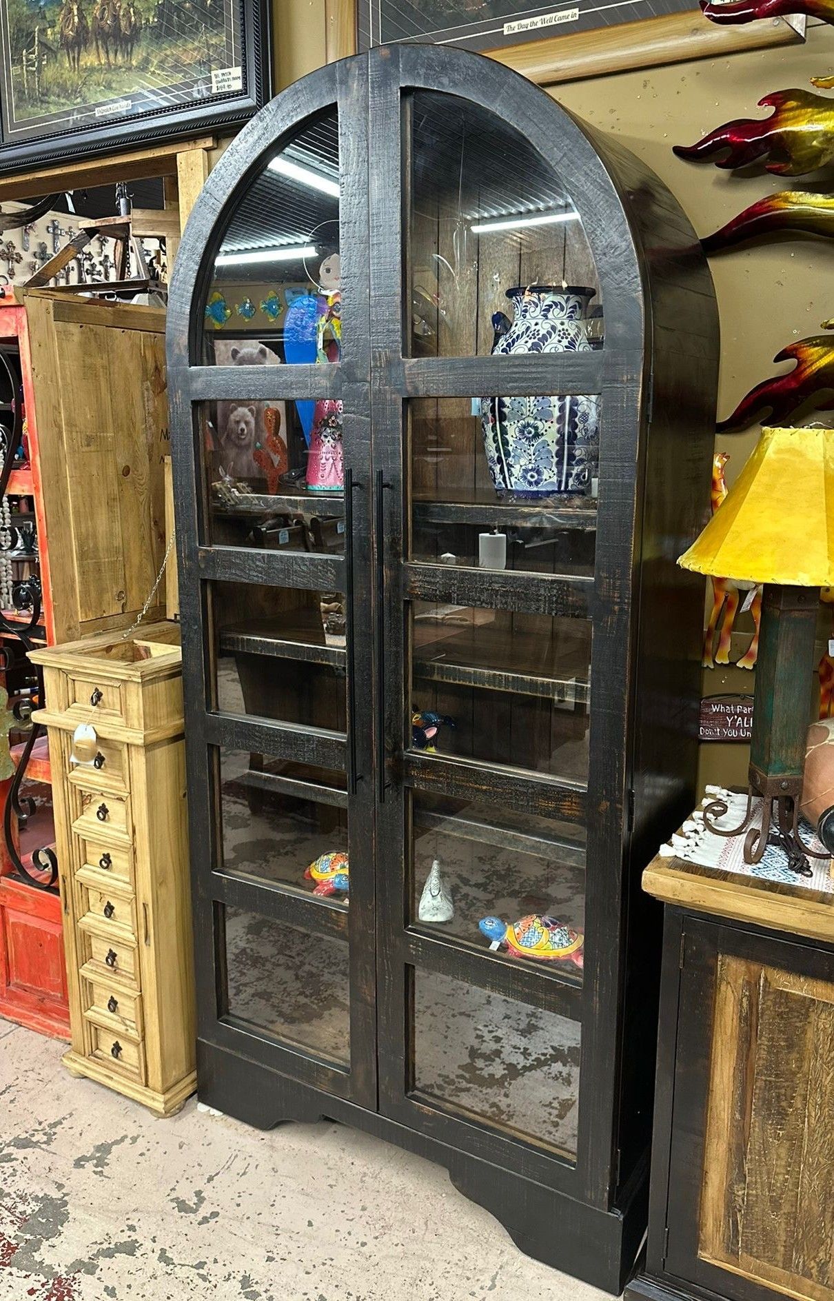 Tall, black cabinet with arched top and glass doors, displaying various items on shelves. The setting is a shop with other furniture and decor.