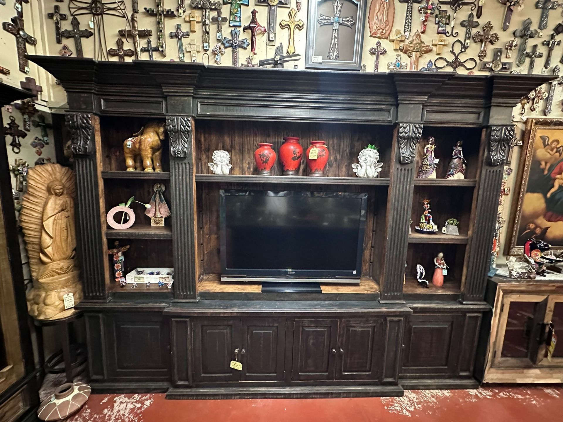 Large, dark wooden entertainment center with a TV, shelves, and religious figurines. A wall of crosses forms the backdrop.