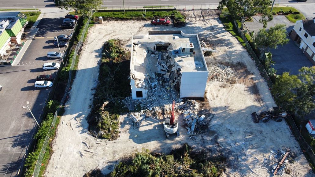 Aerial view of a demolished building site with rubble, a white structure, and surrounding parking lots and trees