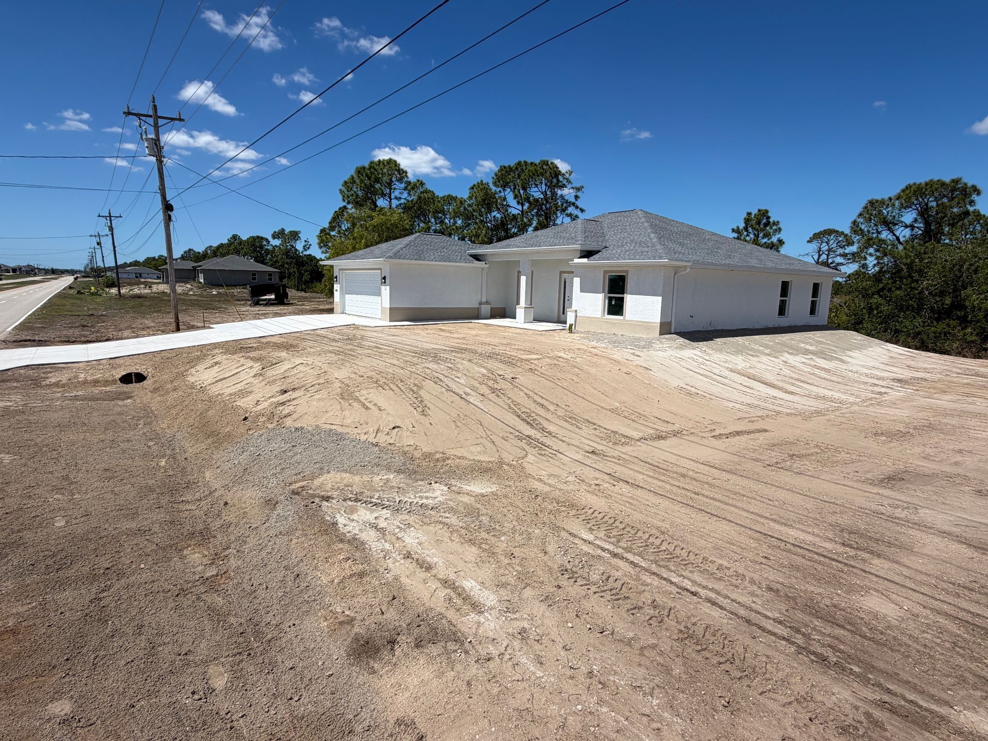 White house under construction with unfinished dirt driveway, utility poles, and trees under a blue sky