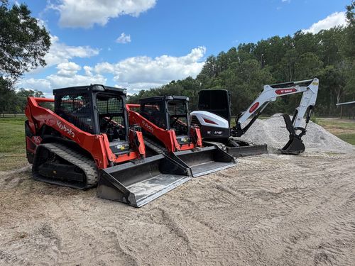 Three compact loaders on a gravel lot under a blue sky, including two red models and a white excavator