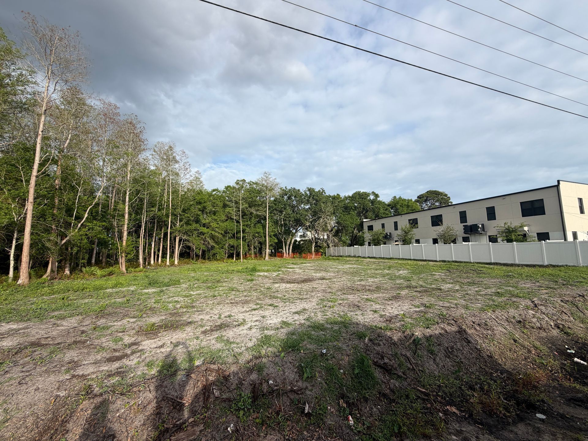 Open grassy lot beside a row of beige townhouses under a cloudy sky, with trees and power lines nearby